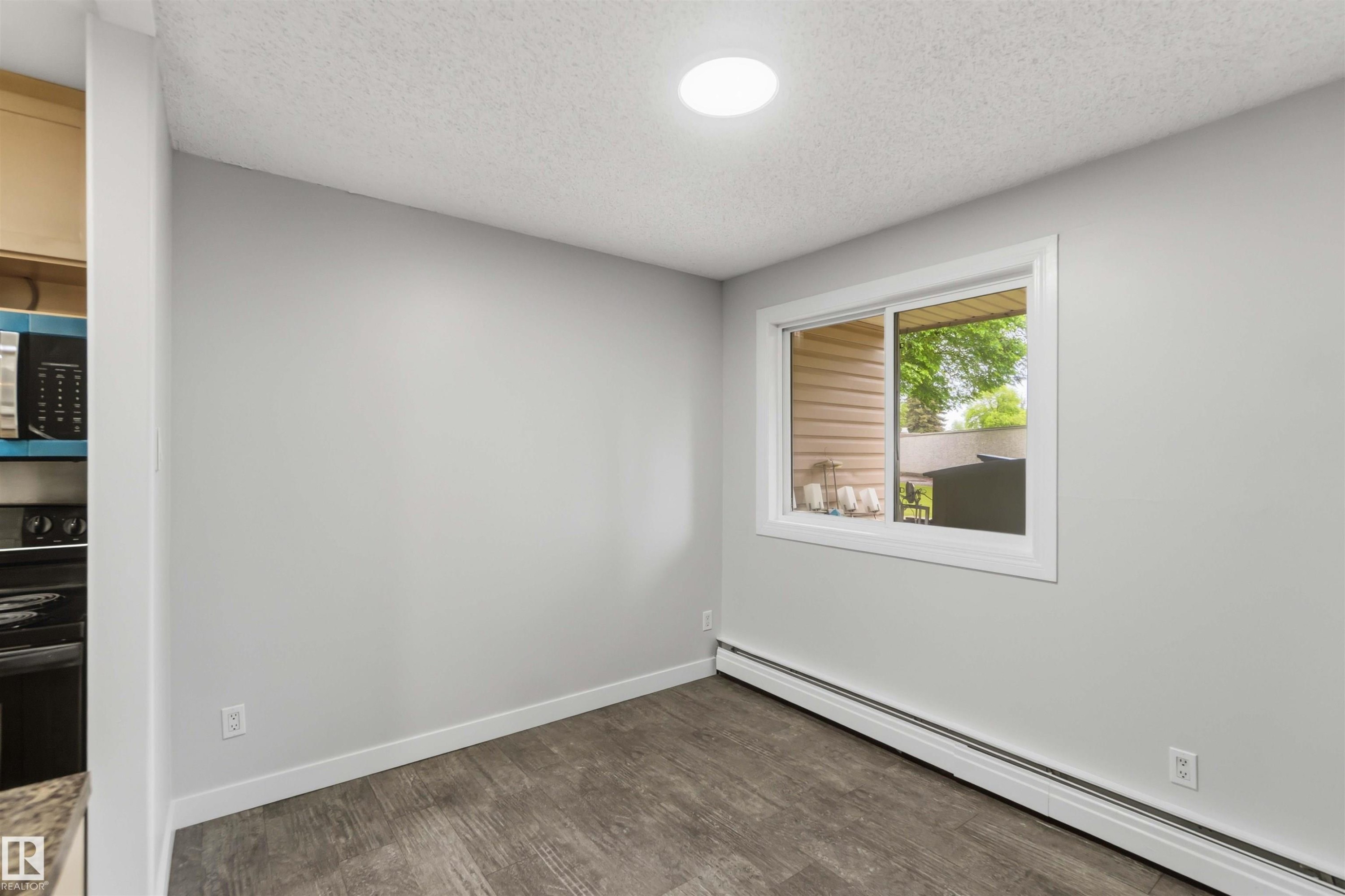 Bright room with wood-finish flooring, featuring a large window, white trim, and a ceiling-mounted light fixture - #121 5125 Riverbend Rd Nw Nw, Edmonton, AB - Indoor Photo Showing Other Room