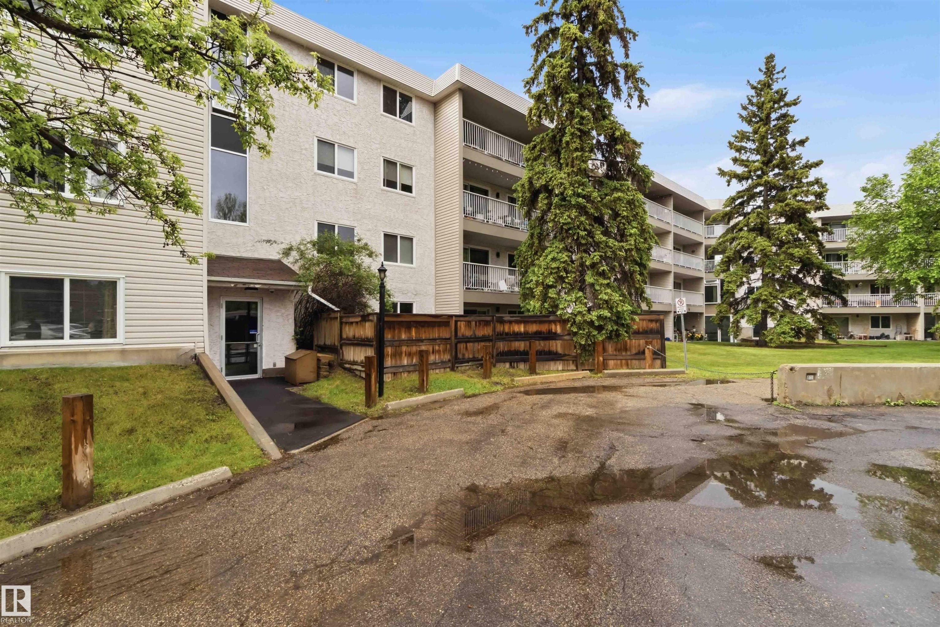Multi-story building featuring balconies with metal railings, a paved parking area, and surrounding green spaces with mature evergreen trees - #121 5125 Riverbend Rd Nw Nw, Edmonton, AB - Outdoor