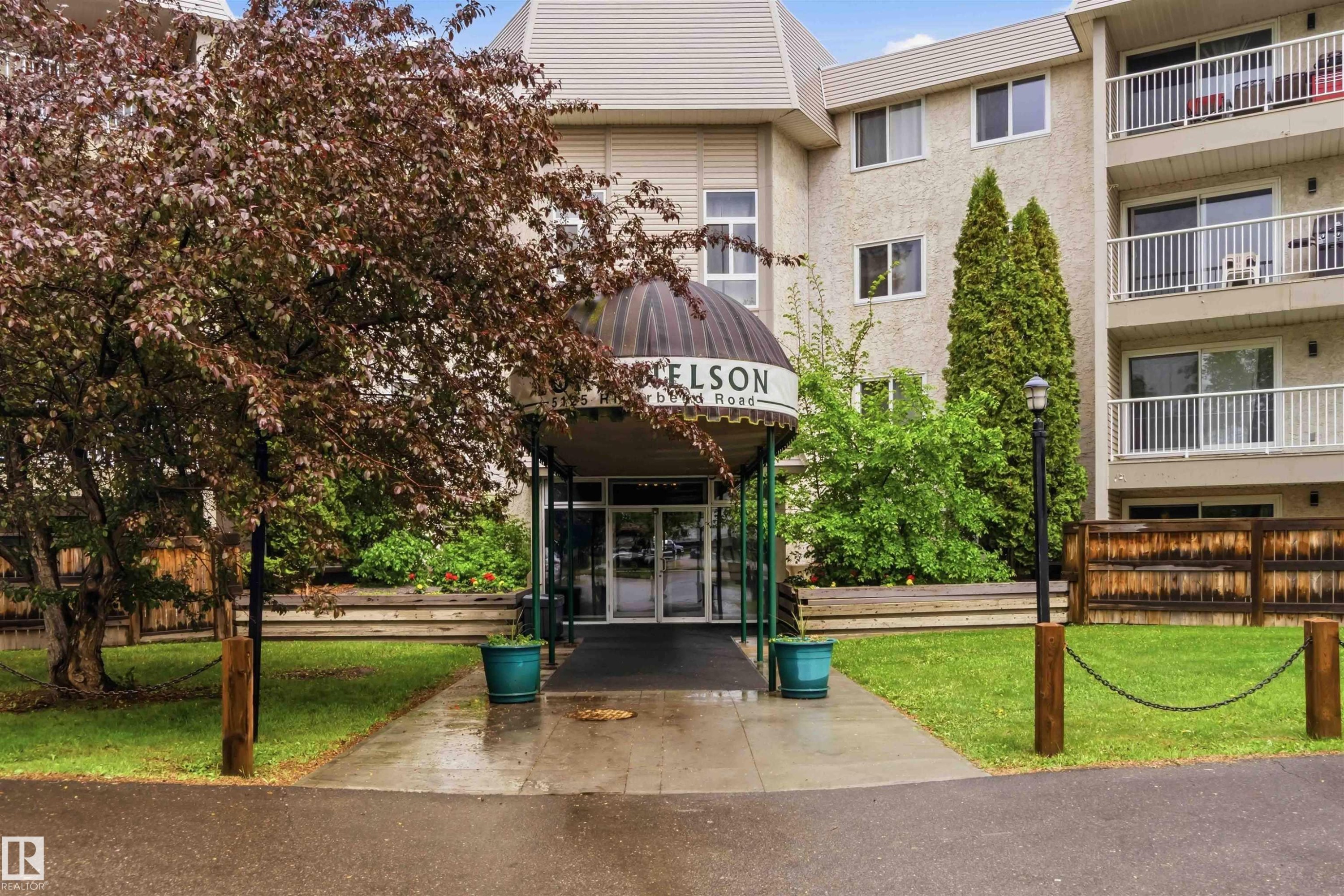Building exterior featuring a covered entrance, multiple balconies with white railings, and a landscaped frontage with mature trees and shrubs - #121 5125 Riverbend Rd Nw Nw, Edmonton, AB - Outdoor