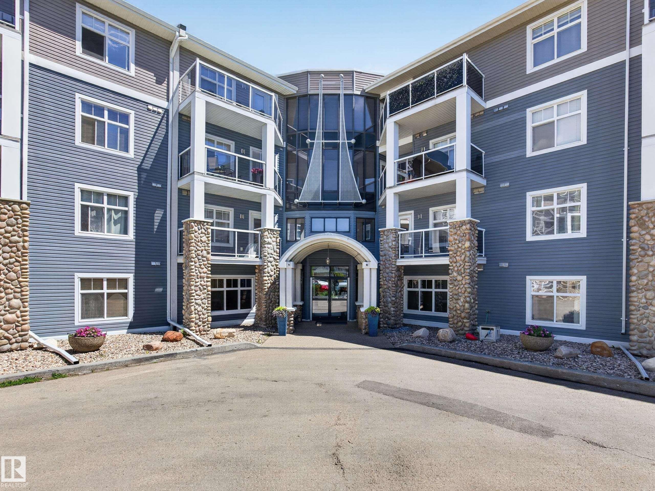Multi-story building with blue siding, stone-clad columns, a central glass facade, multiple balconies with glass railings, and an arched entryway - 138 16035 132 Street, Edmonton, AB