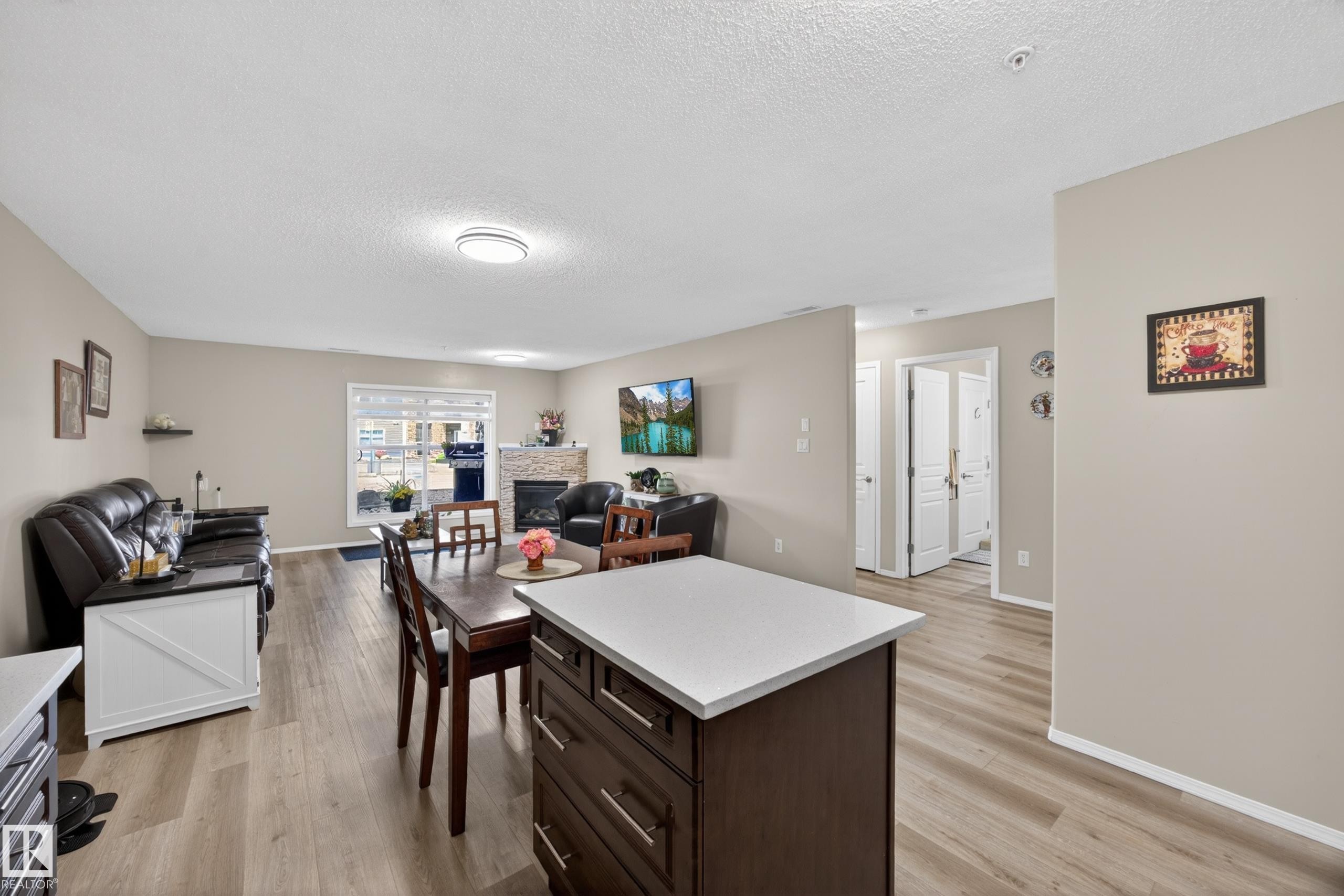Open-concept living area featuring wood-finish flooring, a contemporary kitchen island with a light-colored countertop, and a fireplace with a stone surround - 138 16035 132 Street, Edmonton, AB