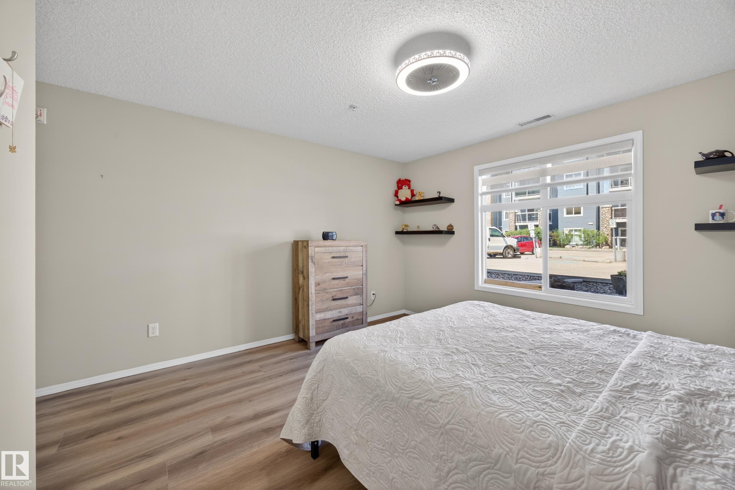 Spacious room featuring wood-finish flooring, a large window with white framing, and a modern ceiling light fixture - 138 16035 132 Street, Edmonton, AB