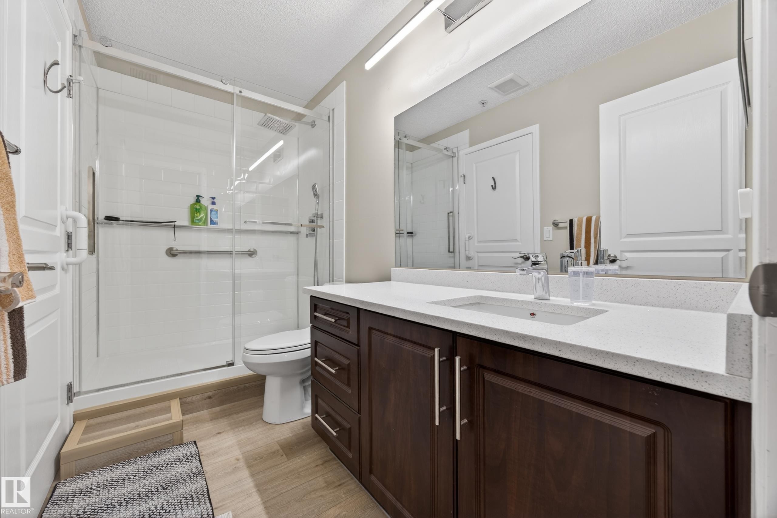 Spacious bathroom featuring a glass-enclosed shower with white subway tile, a dark wood-finish vanity with a light-toned countertop, a rectangular undermount sink, and a large mirror - 138 16035 132 Street, Edmonton, AB