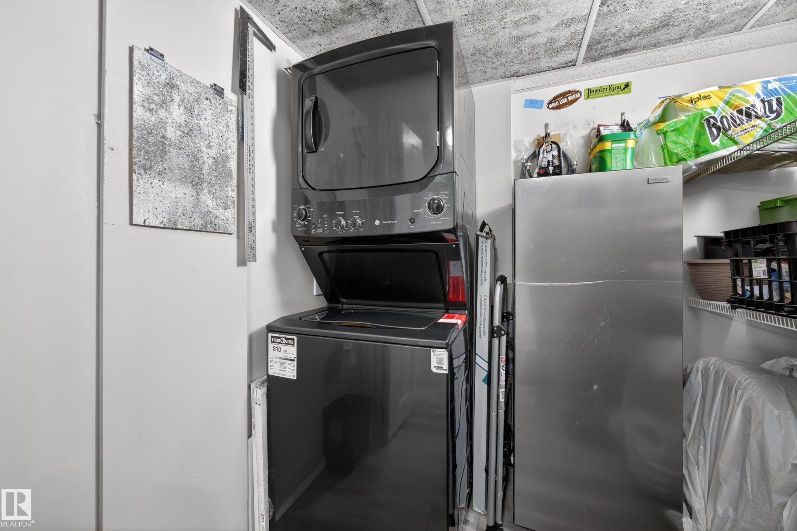 Compact laundry area featuring a stacked washer and dryer, adjacent to a stainless steel refrigerator and built-in shelving - 138 16035 132 Street, Edmonton, AB