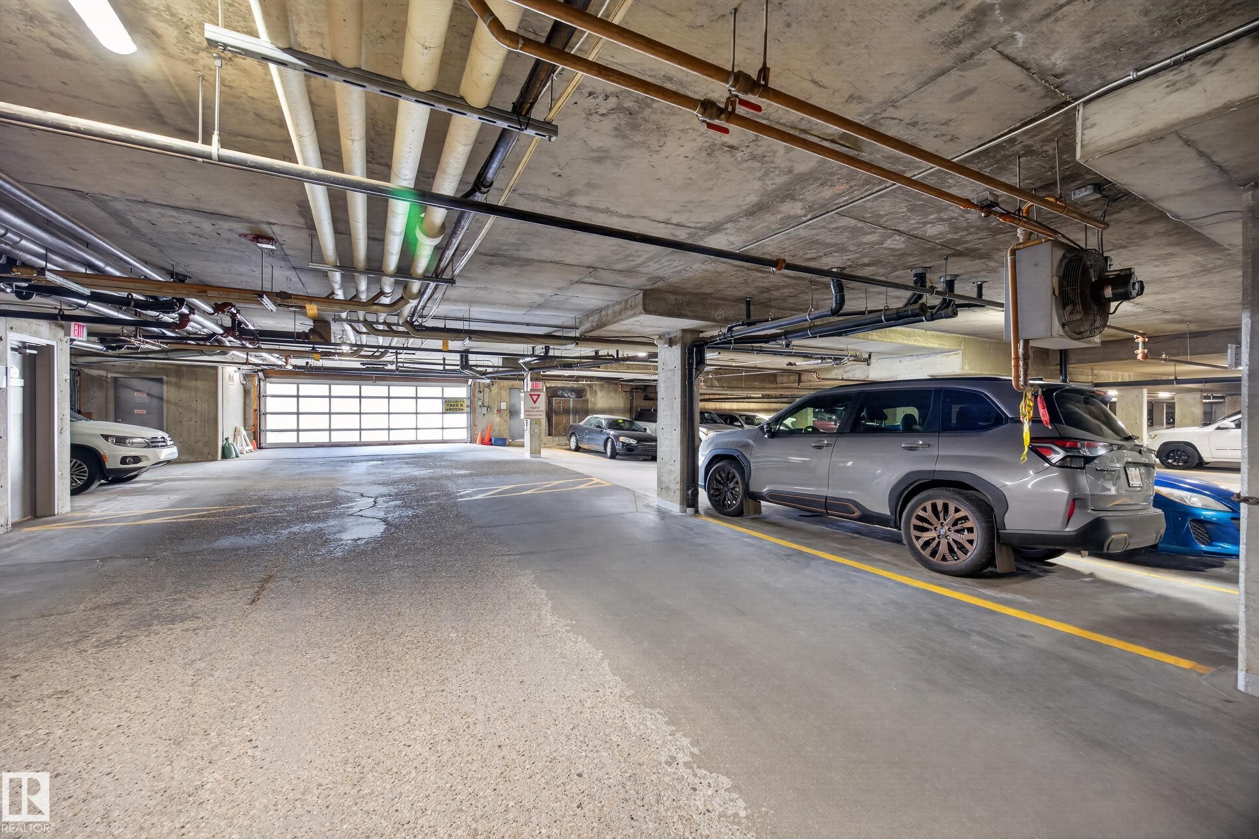 Underground parking garage featuring exposed concrete ceiling, overhead utility piping, and fluorescent lighting - 138 16035 132 Street, Edmonton, AB