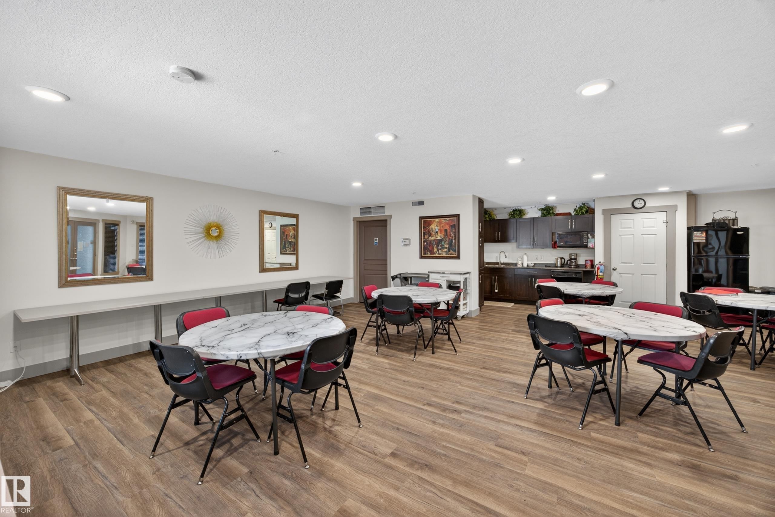 Spacious common room featuring wood-finish flooring, recessed lighting, and a built-in kitchen area with dark cabinetry and a microwave - 138 16035 132 Street, Edmonton, AB