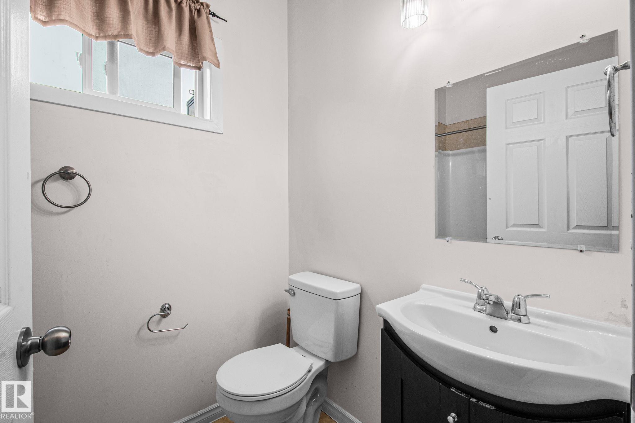 Bathroom featuring a window, white toilet, dark vanity with integrated sink, wall-mounted mirror, and overhead light fixture - 4806 117 Ave, Edmonton, AB