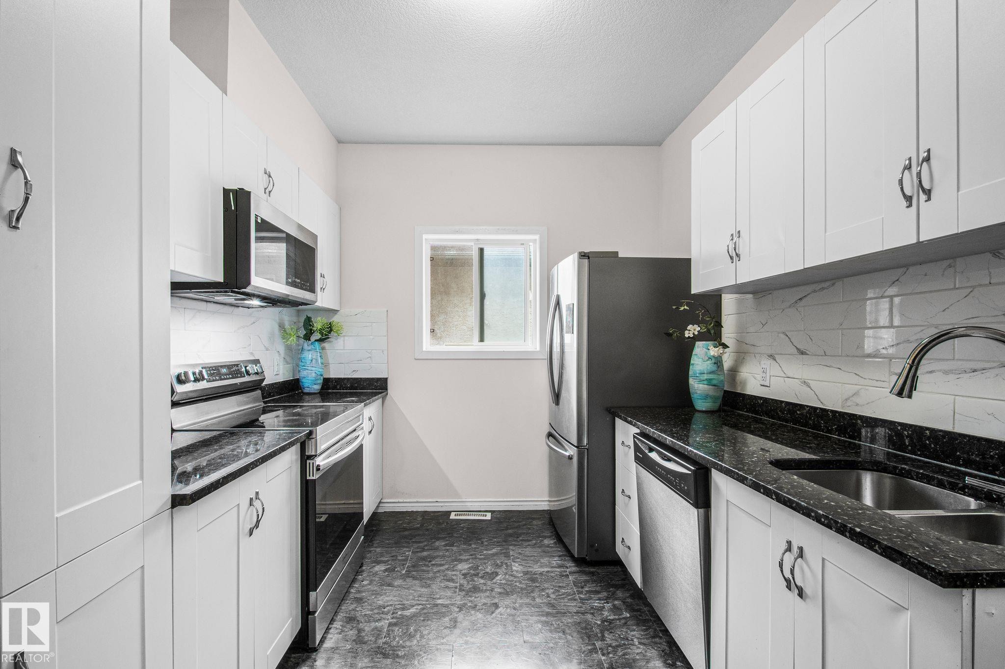 Galley kitchen featuring white cabinetry, dark speckled countertops, stainless steel appliances, a white subway tile backsplash, and a window - 4806 117 Ave, Edmonton, AB