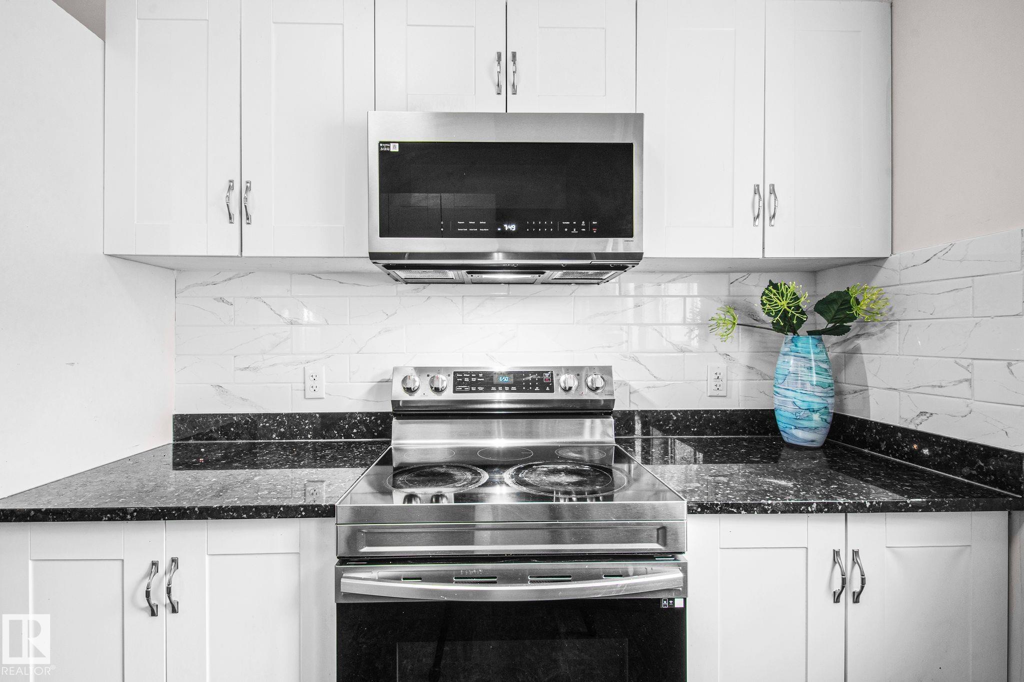 Modern kitchen featuring a stainless steel range with overhead microwave, white cabinetry with silver hardware, black speckled countertops, and a white subway tile backsplash - 4806 117 Ave, Edmonton, AB