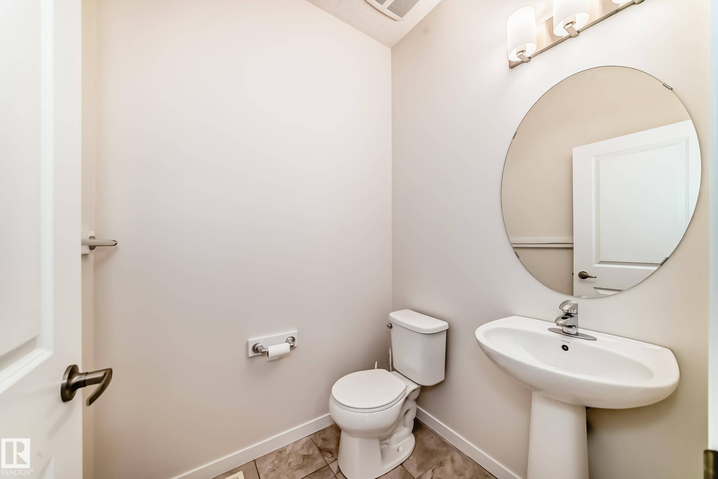 Pedestal sink with a circular mirror, brushed nickel light fixture, toilet, and tile flooring - 18103 77 Street, Edmonton, AB