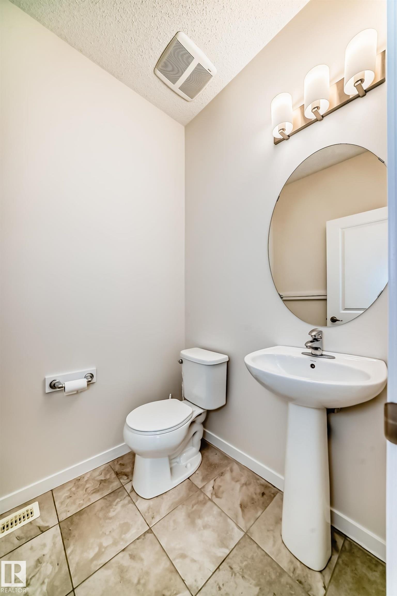 Powder room featuring a pedestal sink, round wall mirror, and a three-light vanity fixture - 18103 77 Street, Edmonton, AB