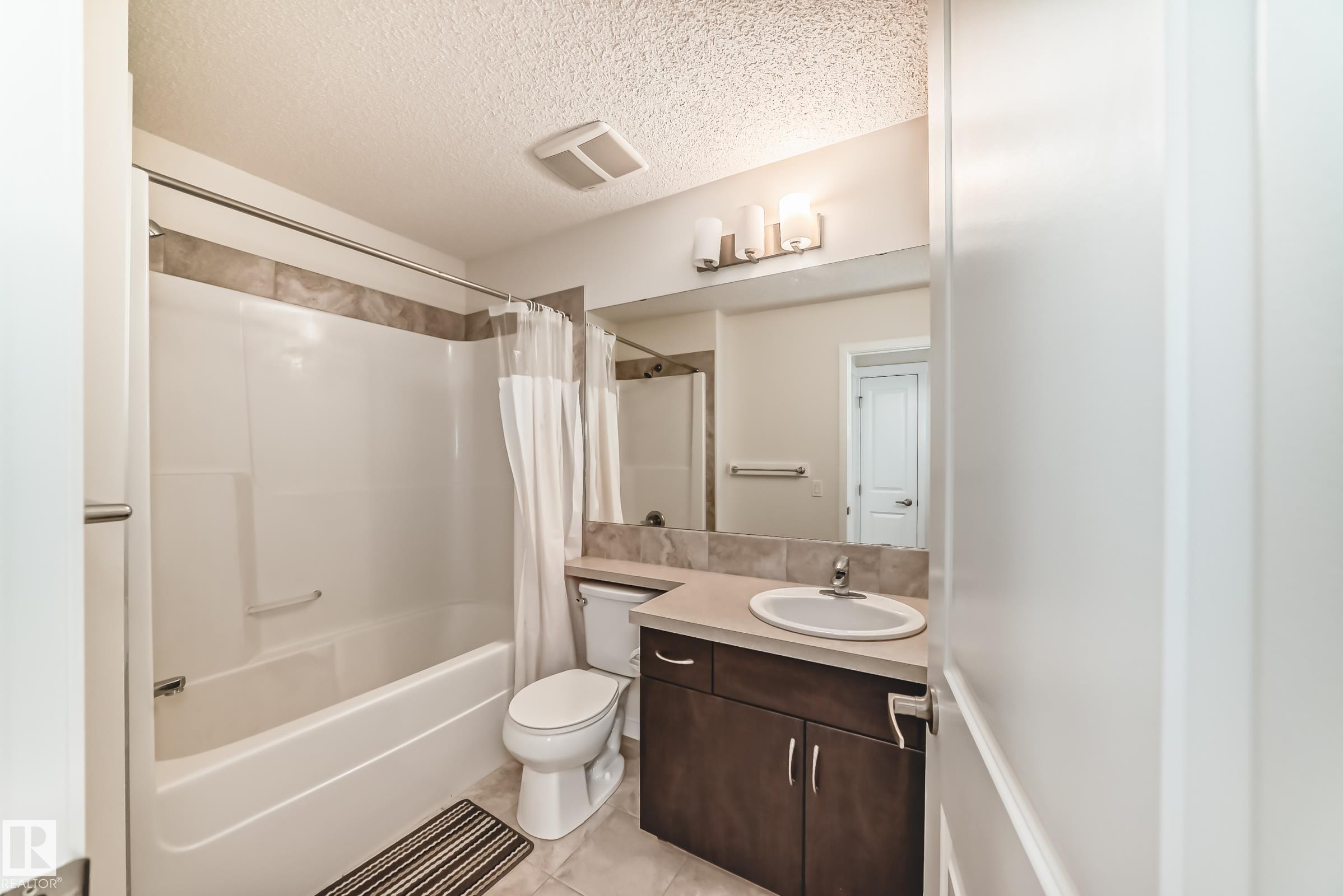 Bathroom featuring a combined tub and shower with tile surround, a dark wood-finish vanity with an integrated sink and light-toned countertop, and an overhead vanity light fixture - 18103 77 Street, Edmonton, AB