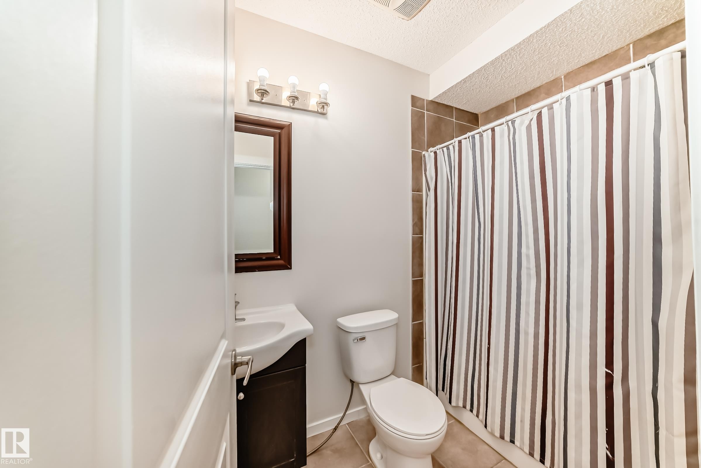 Bathroom featuring ceramic tile flooring, a dark wood-framed mirror, a vanity with a white sink, and a tiled shower surround - 18103 77 Street, Edmonton, AB
