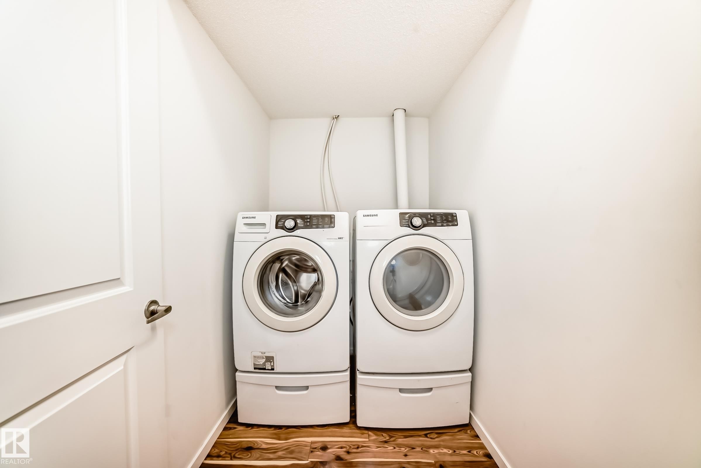 Dedicated laundry area featuring a front-loading washer and dryer set with pedestal bases, wood-finish flooring, and crisp white walls - 18103 77 Street, Edmonton, AB