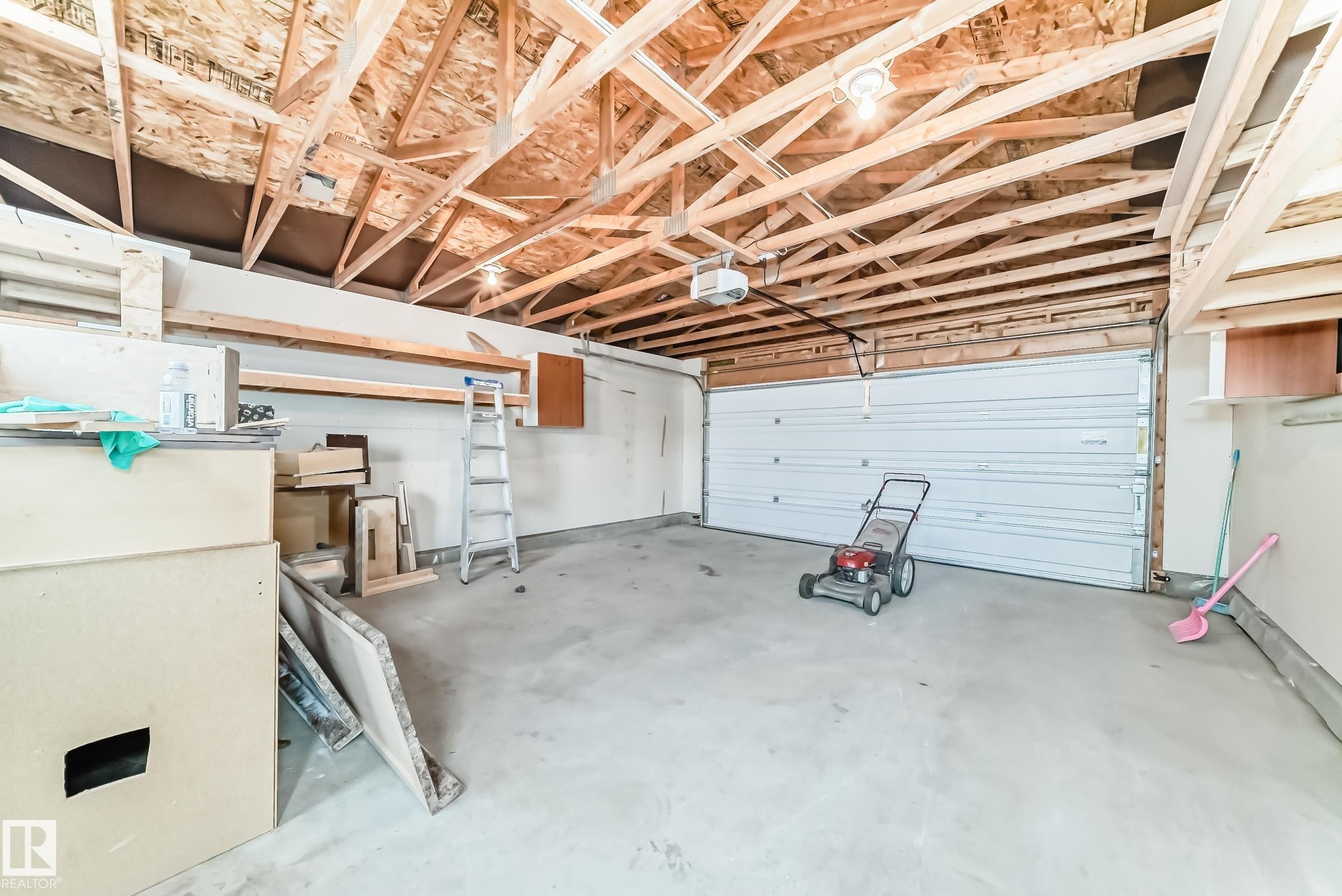 Spacious garage featuring exposed wood truss ceiling, a concrete floor, overhead lighting, and a white panel garage door - 18103 77 Street, Edmonton, AB