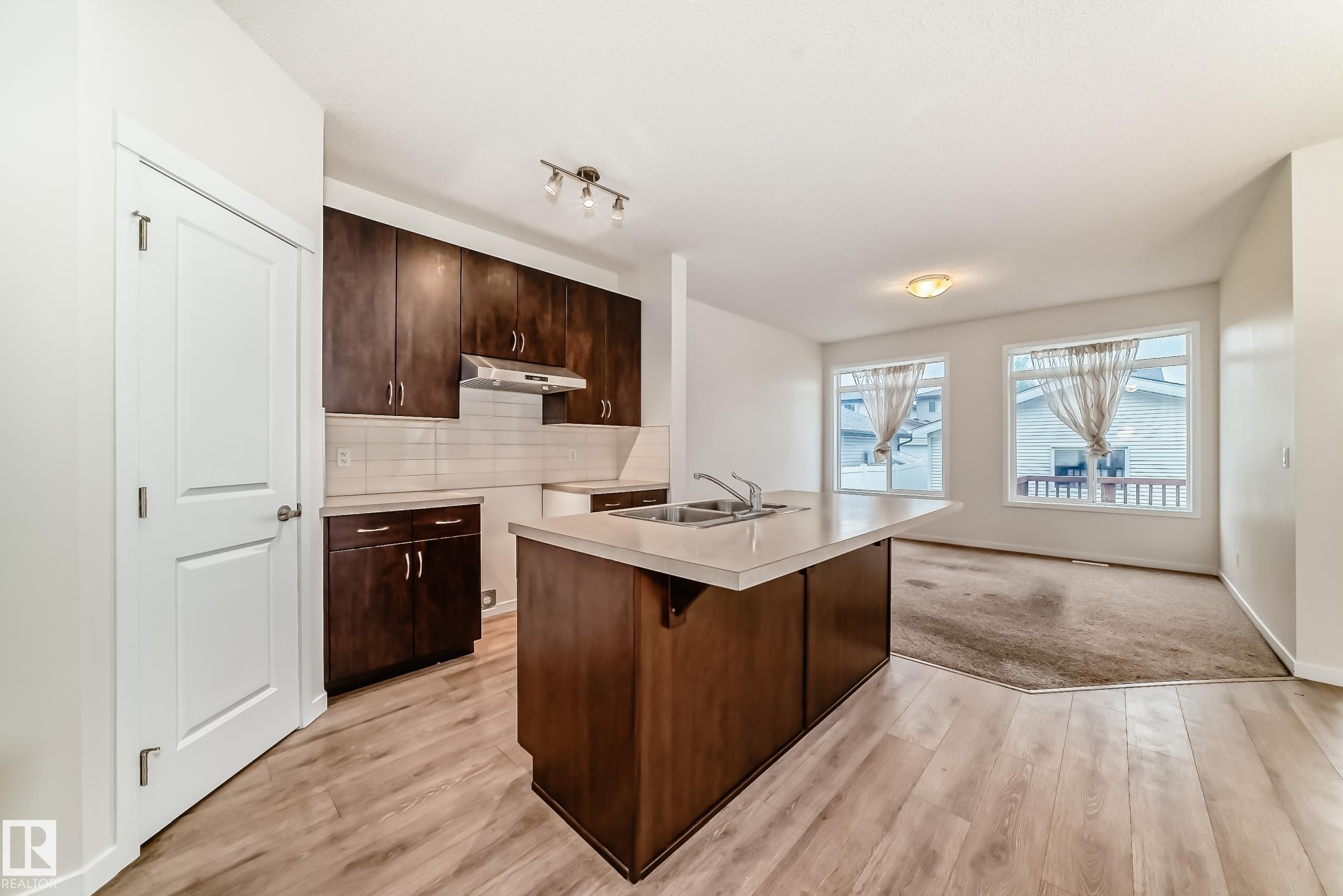 Open-concept kitchen featuring wood-finish flooring, dark wood cabinetry, a white tile backsplash, a kitchen island with a double sink, and track lighting - 18103 77 Street, Edmonton, AB