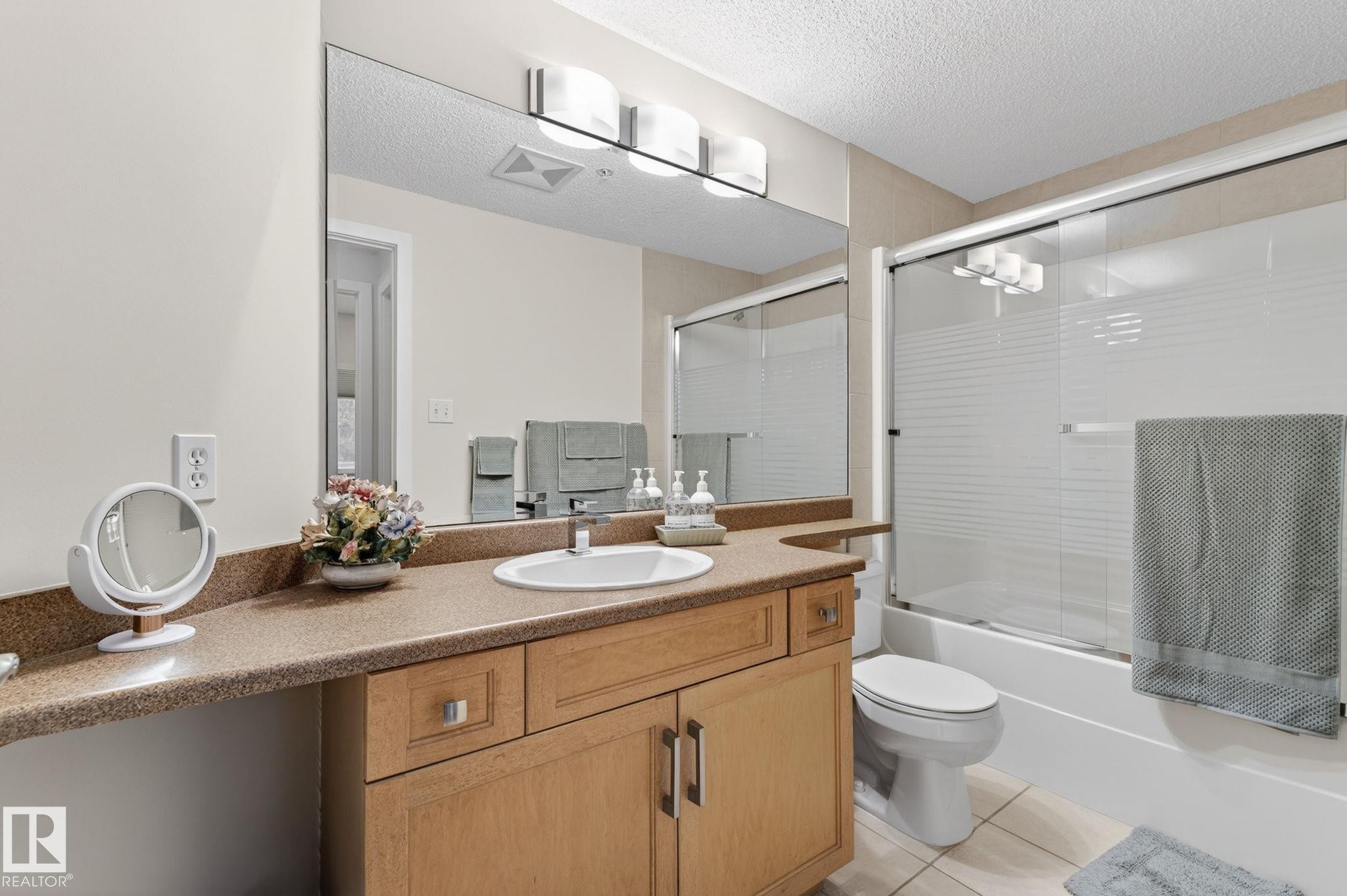 Bathroom featuring a single vanity with a white basin and stone-finish countertop, light wood-tone cabinetry, a large wall-mounted mirror, and a shower-tub combination with a glass enclosure - 208 5 Gate Avenue, St. Albert, AB