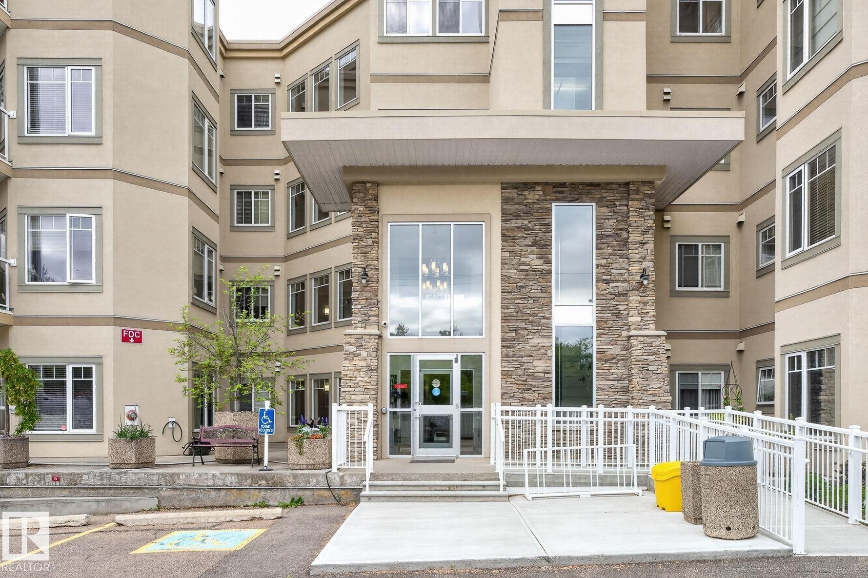 Multi-story building featuring a stone-clad entrance with large glass doors and windows, a covered entryway, and a concrete accessibility ramp with white railings - 208 5 Gate Avenue, St. Albert, AB