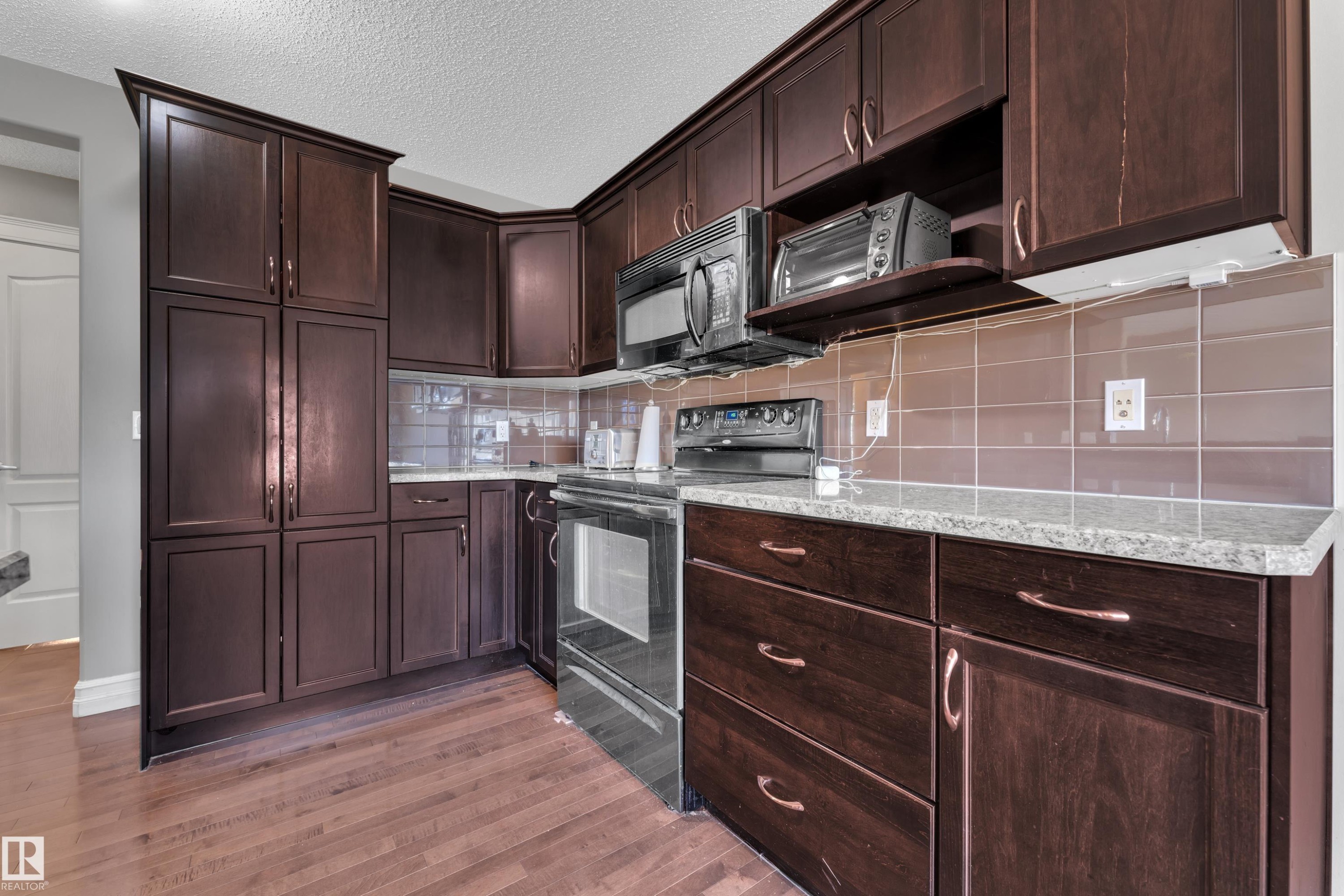 Kitchen featuring extensive dark wood-finish cabinetry, light-toned countertops, a tile backsplash, stainless steel appliances, and wood-finish flooring - 6116 11 Avenue, Edmonton, AB