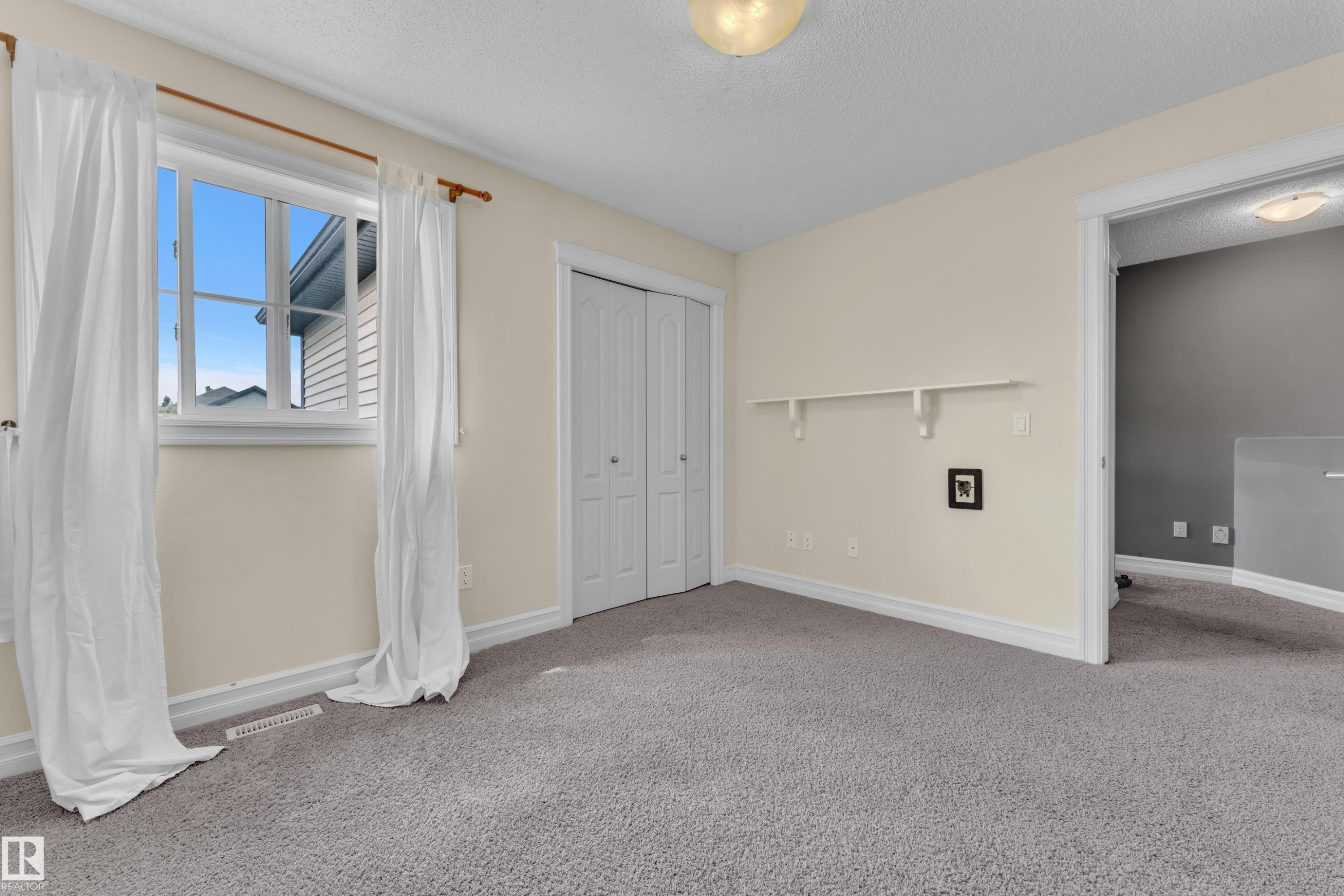 Carpeted interior space featuring a window with white trim, bi-fold closet doors, a built-in wall shelf, and a ceiling-mounted light fixture - 6116 11 Avenue, Edmonton, AB