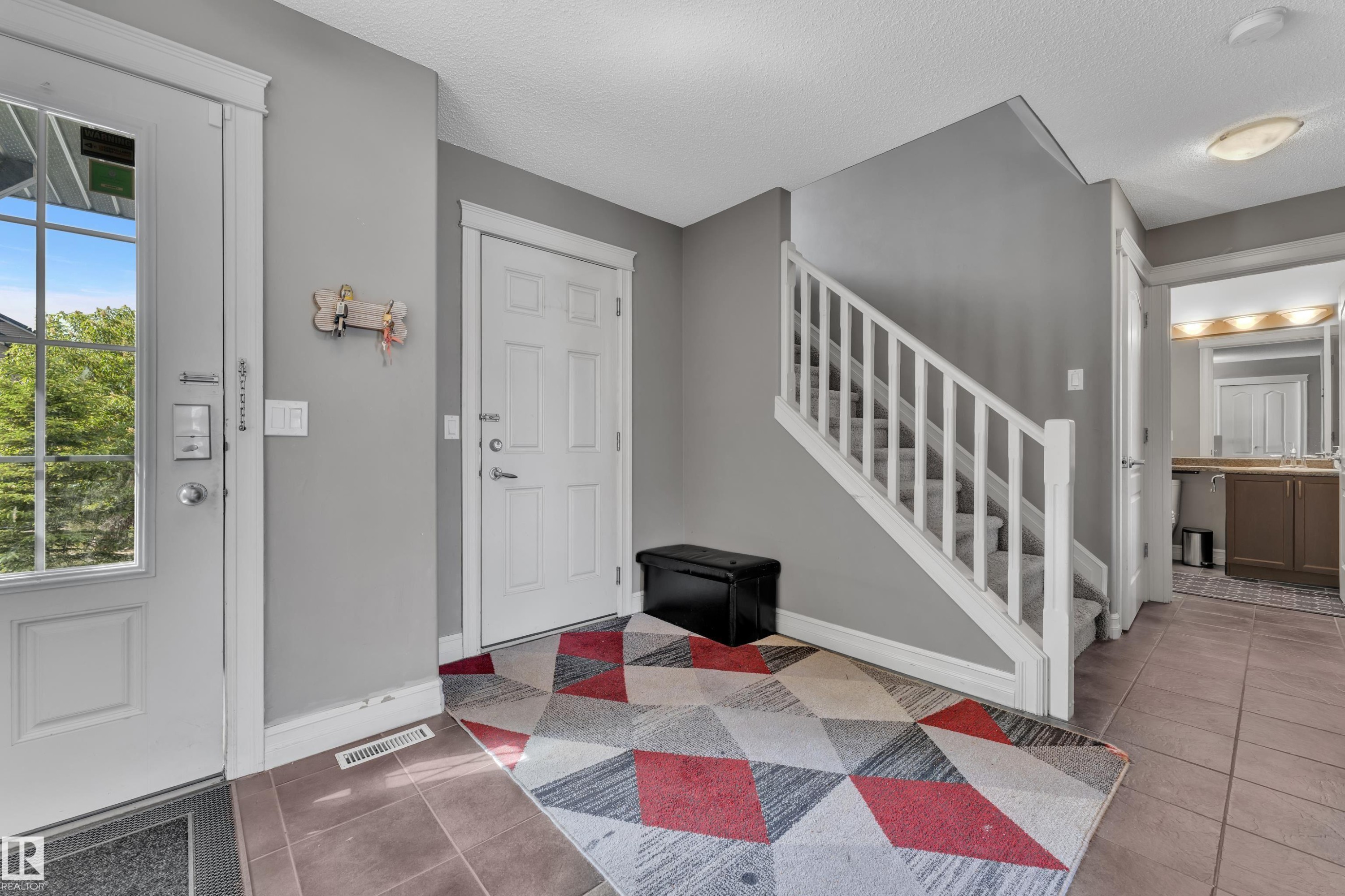 Entrance foyer featuring ceramic tile flooring, a staircase with carpeted treads and white wood railing, a partial light gray painted wall, and a powder room with a vanity and mirror - 6116 11 Avenue, Edmonton, AB