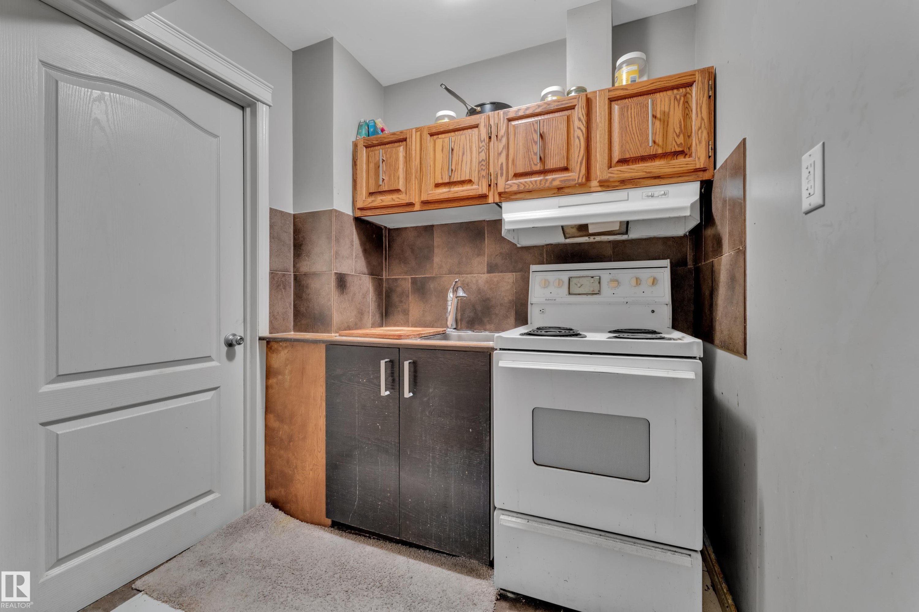 Compact kitchen area featuring a white electric range, integrated range hood, and a stainless steel sink with a chrome faucet - 6116 11 Avenue, Edmonton, AB