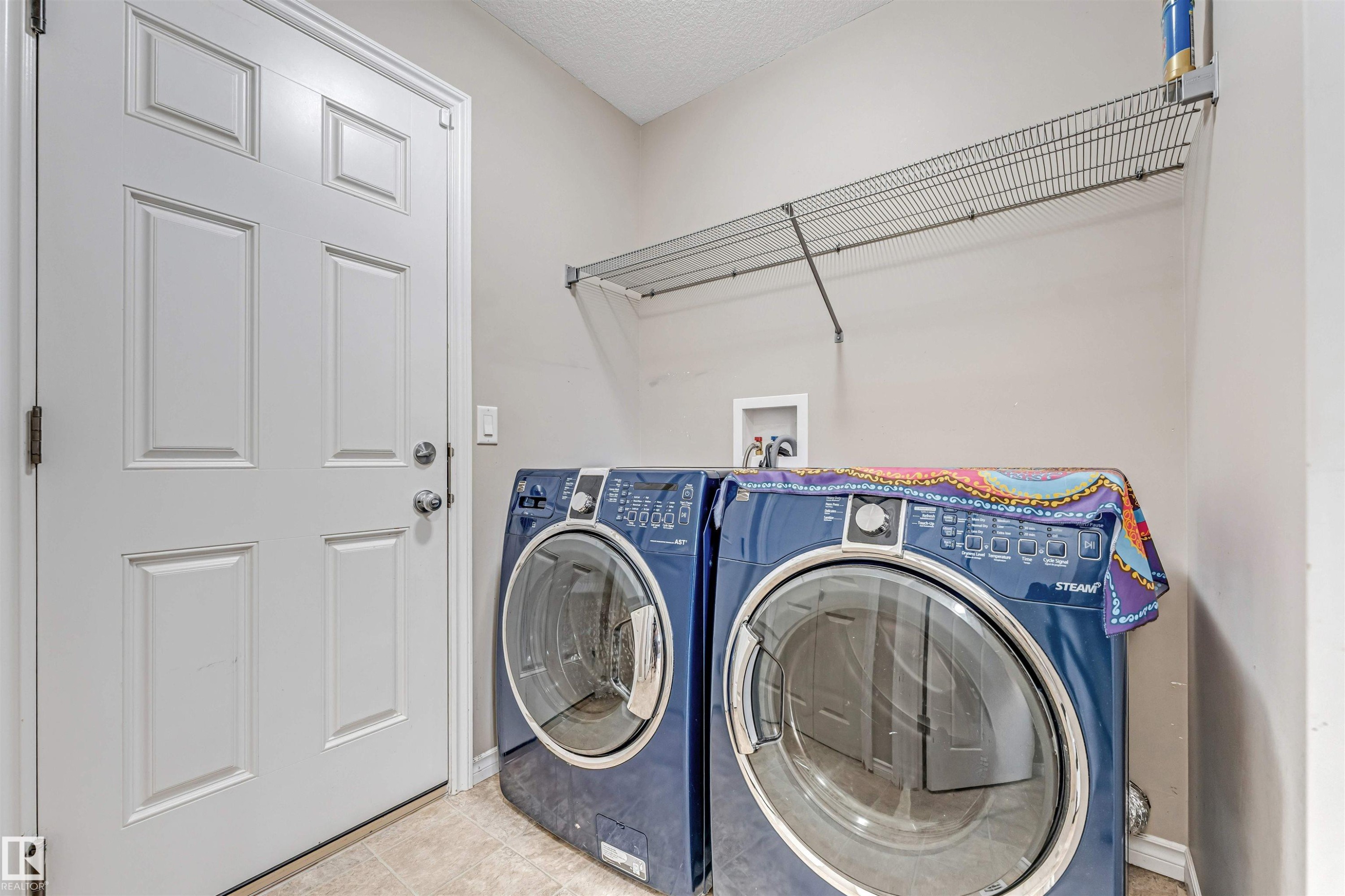 Dedicated laundry area featuring a white paneled door, light-toned wall paint, and overhead wire shelving - 5928 12 Avenue, Edmonton, AB