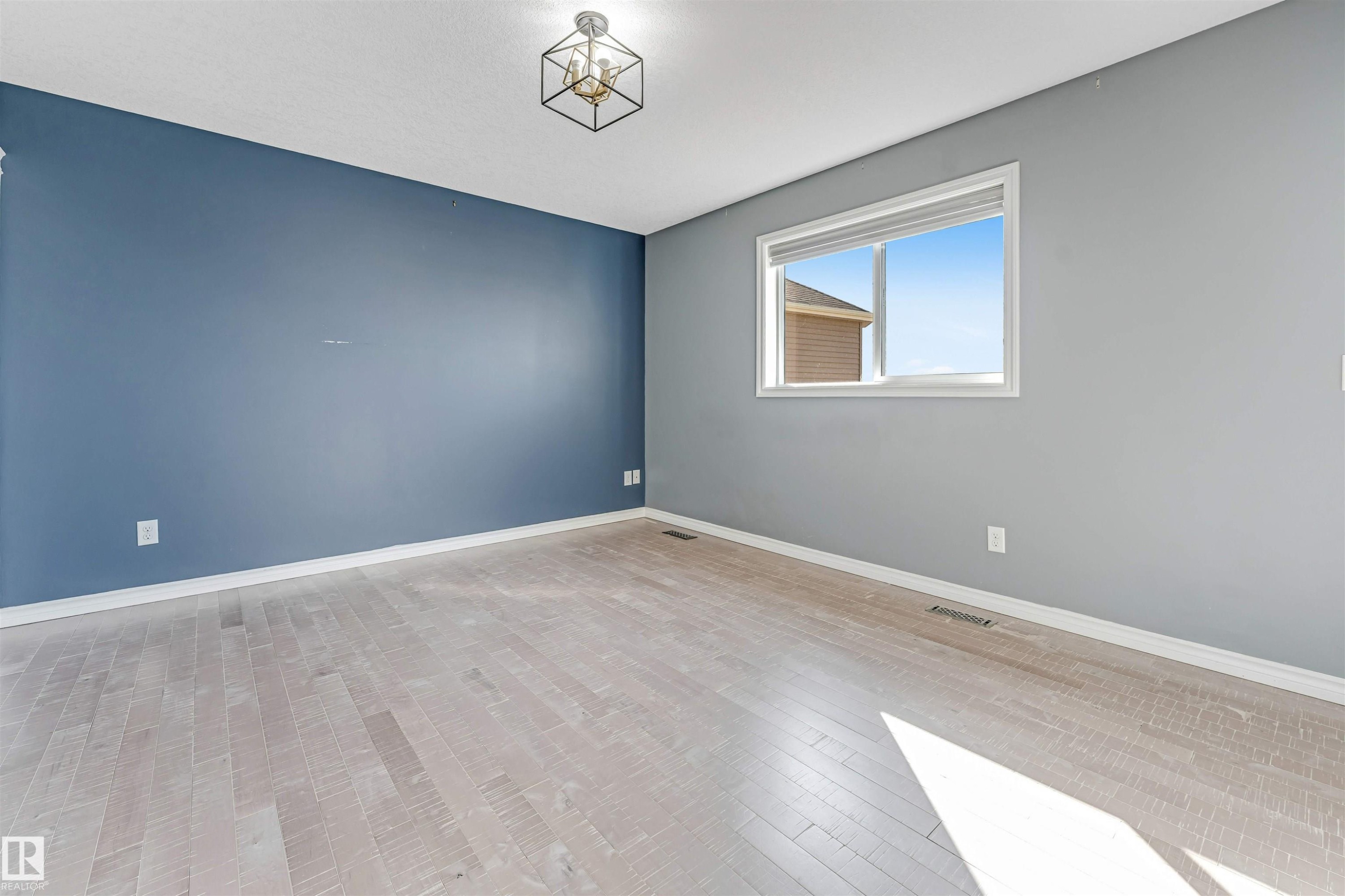 Spacious room featuring light wood-finish flooring, a contemporary ceiling light fixture, and a large window with white trim - 5928 12 Avenue, Edmonton, AB