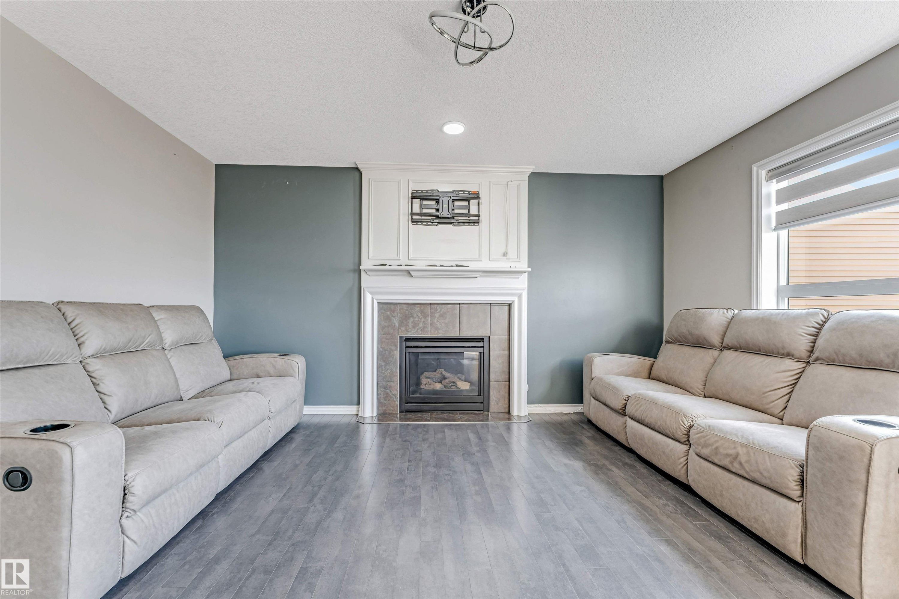Living area featuring a central fireplace with a tile surround and white mantelpiece, recessed lighting, wood-finish flooring, and a large window with roller blinds - 5928 12 Avenue, Edmonton, AB