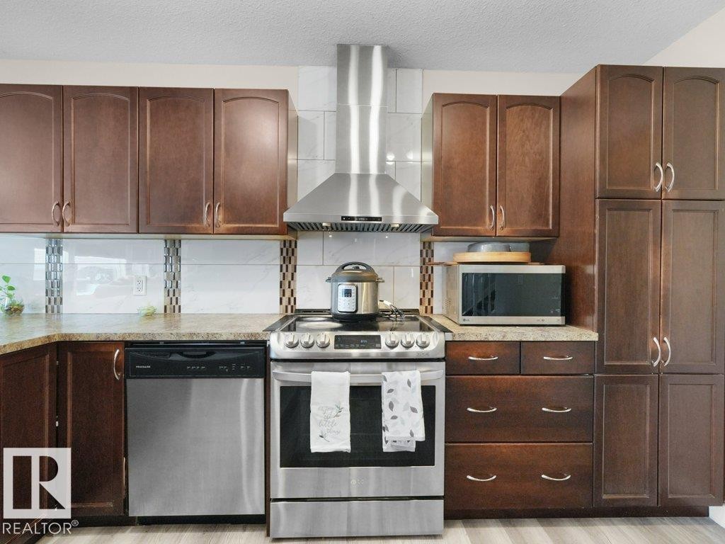 Stainless steel range and matching range hood, brown wood-finish cabinetry, light-toned countertops, tile backsplash, and light wood-finish flooring - 3756 8 Street, Edmonton, AB