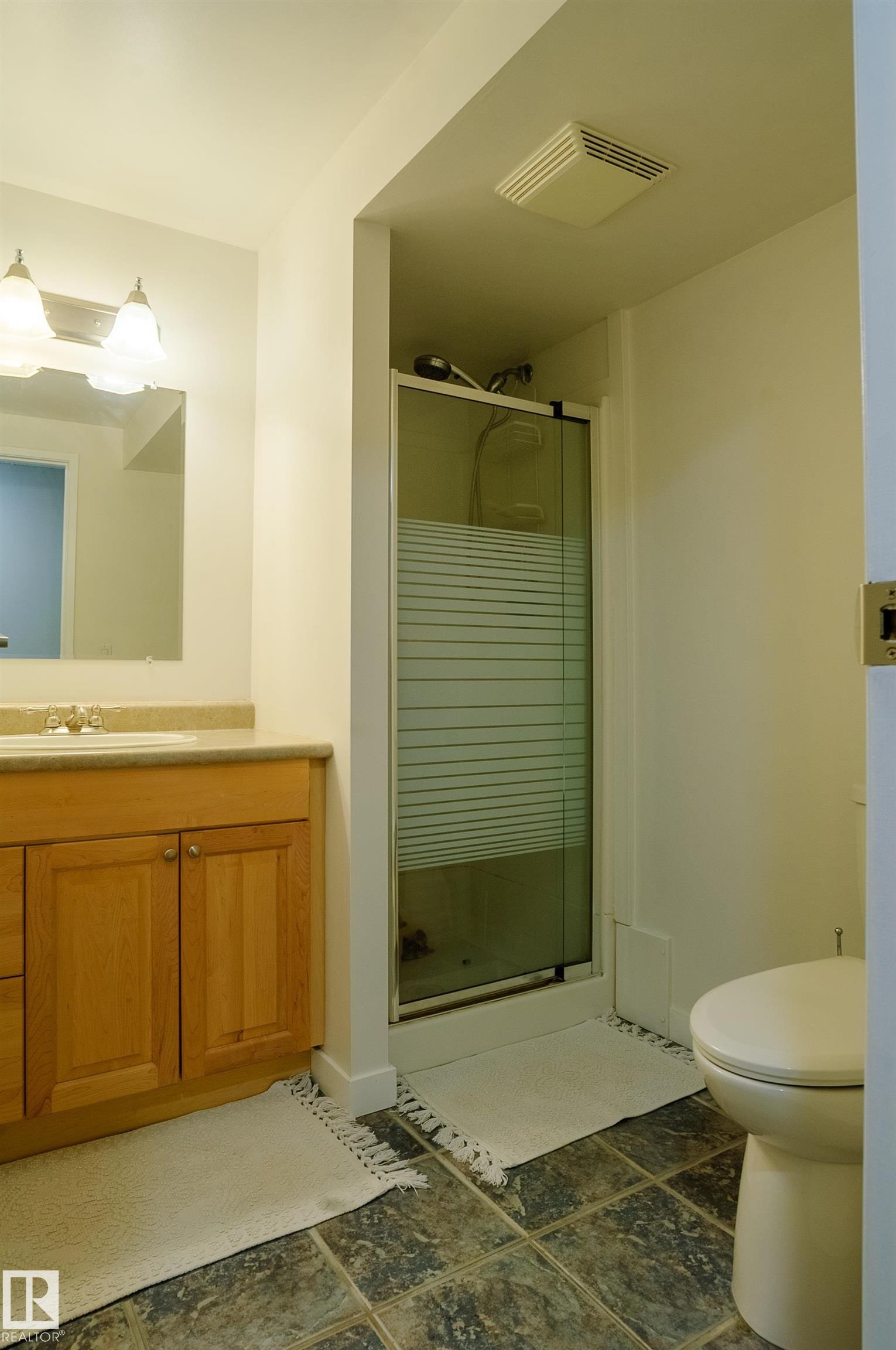 Basement bathroom featuring a glass-enclosed shower with frosted paneling, a single vanity with wood-finish cabinetry, a white ceramic toilet, and square-tiled flooring - 58 Lodgepole Crescent, St. Albert, AB