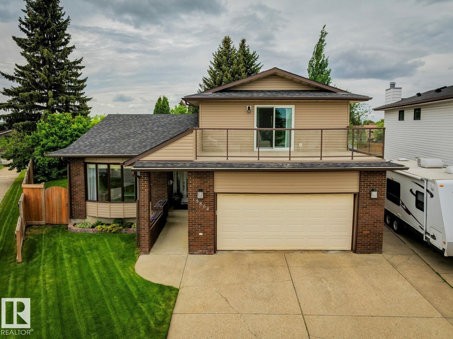 Two-story residence featuring a ground-level garage, second-story balcony with glass railing, bay window, and a fenced yard with mature trees - 15804 80 Street, Edmonton, AB