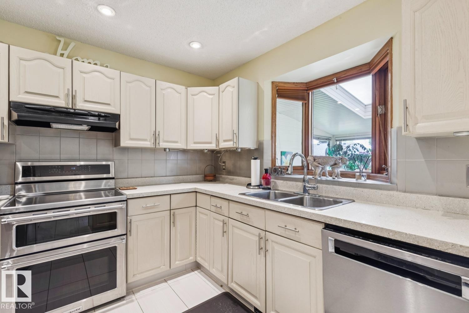 Kitchen featuring cream cabinetry, light-toned countertops, a double basin stainless steel sink, and a multi-pane window with a wood-finish frame - 15804 80 Street, Edmonton, AB