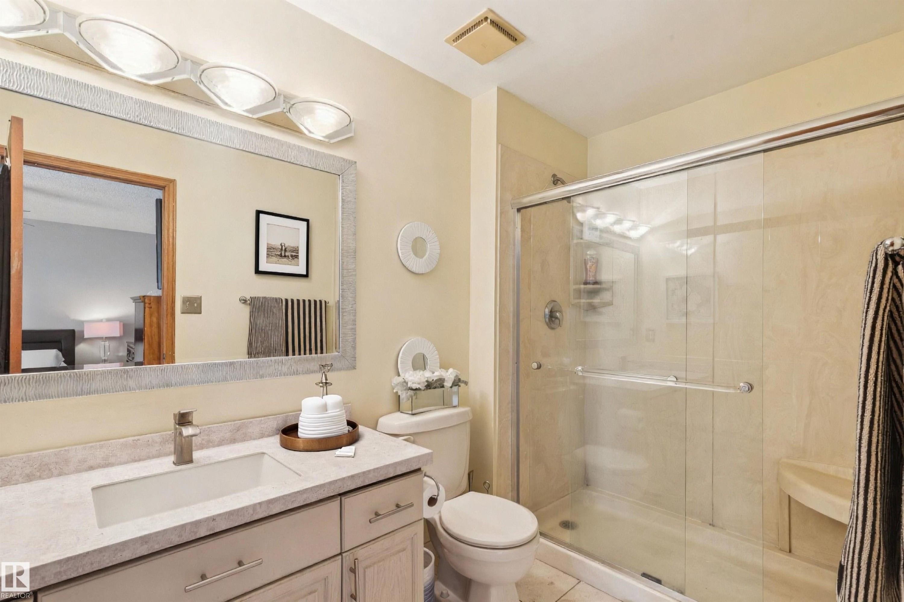 Full primary-bathroom featuring a single vanity with an integrated sink, a large mirror with a textured frame, a toilet, and a glass-enclosed shower with a built-in bench - 15804 80 Street, Edmonton, AB