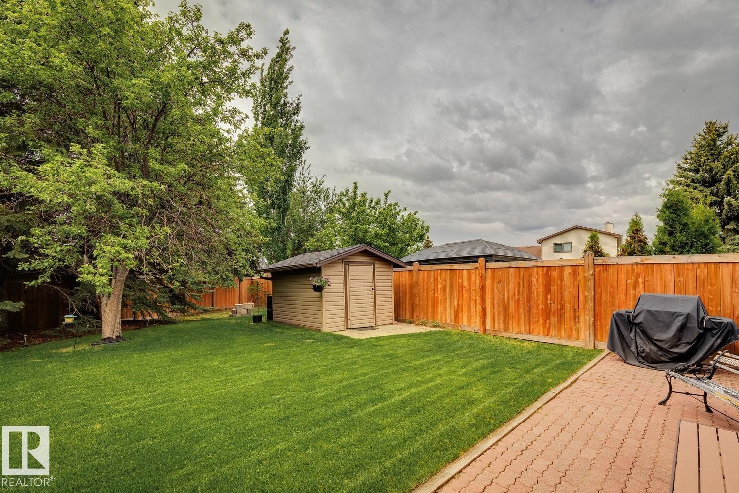 Expansive backyard featuring a manicured lawn, full-grown trees, a tan storage shed, a wood privacy fence, and a paver patio - 15804 80 Street, Edmonton, AB
