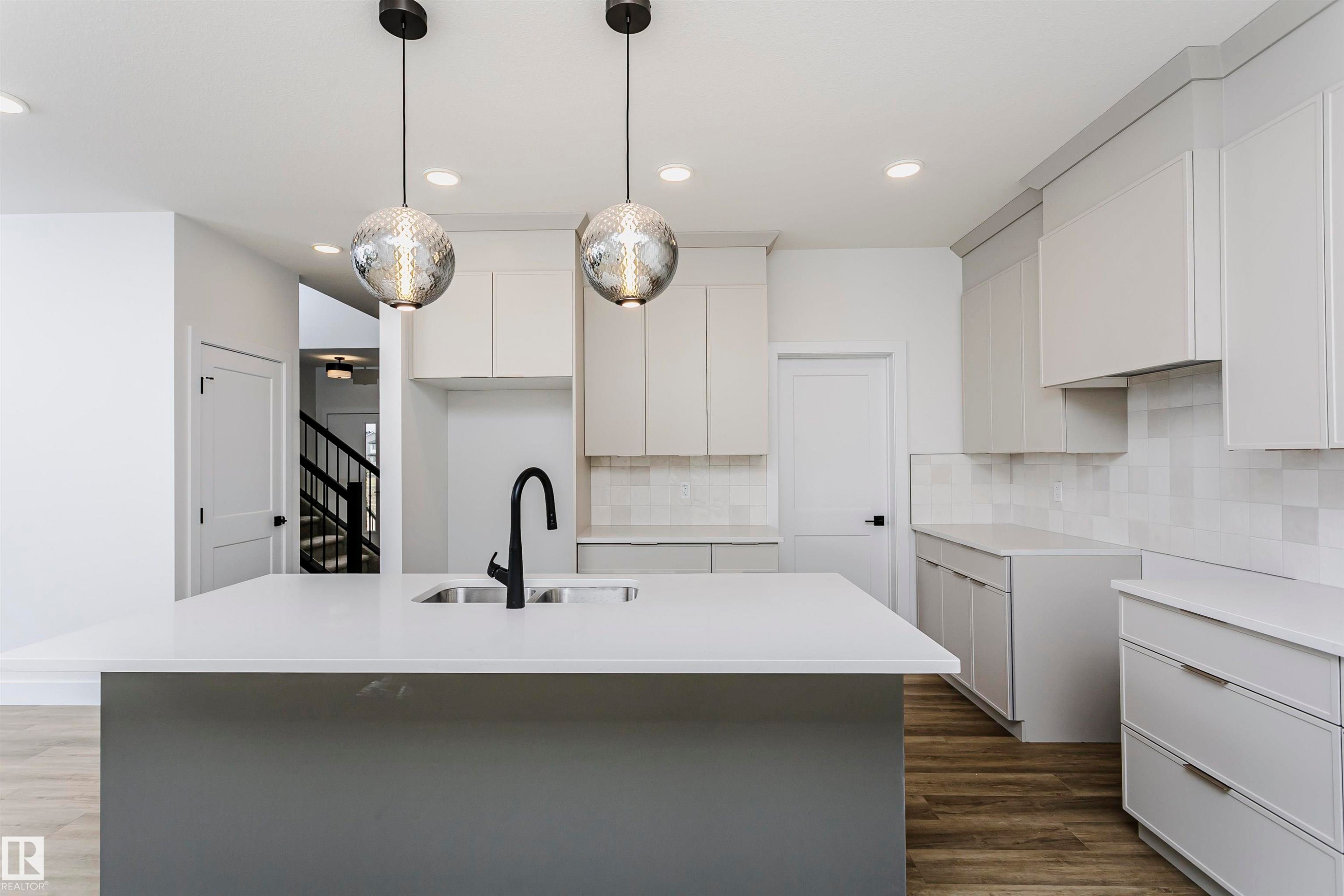 Modern kitchen featuring a central island with an undermount sink and matte black faucet, light grey flat-panel cabinetry, white countertops, and a white tile backsplash - 6210 Dansereau Crescent, Beaumont, AB