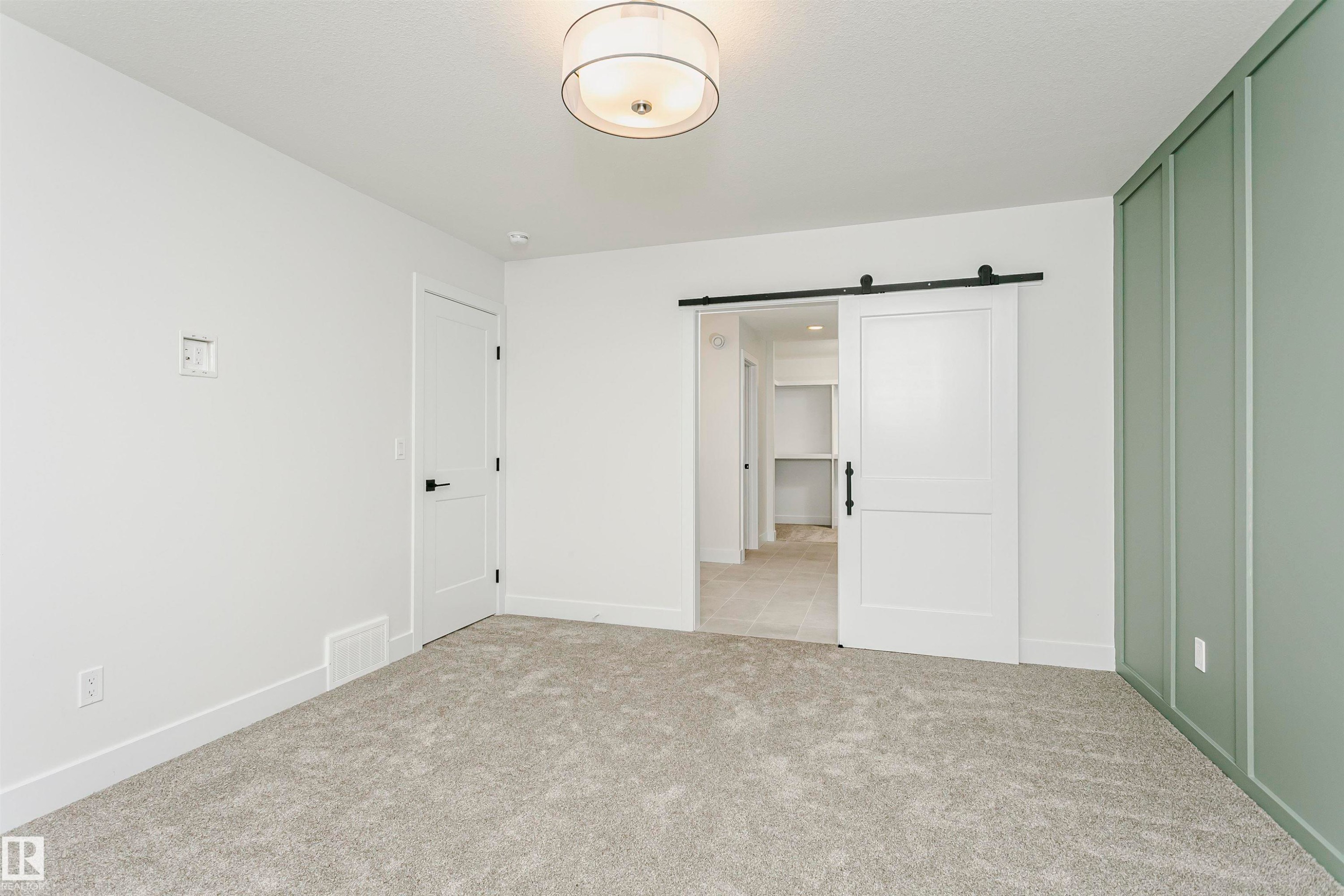 Spacious room featuring light gray carpeting, a decorative wall panel in a soft green hue, and a white barn door on a black rail system - 6210 Dansereau Crescent, Beaumont, AB