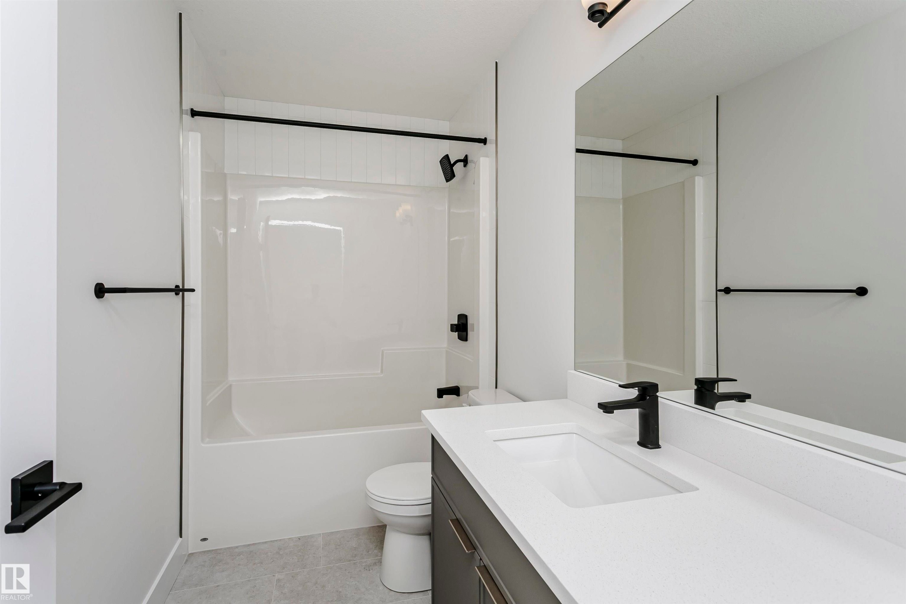 Bathroom featuring a white bathtub and shower combination, dark bronze fixtures, a rectangular vanity mirror, a single-sink vanity with a light-toned countertop, and light gray tile flooring - 6210 Dansereau Crescent, Beaumont, AB