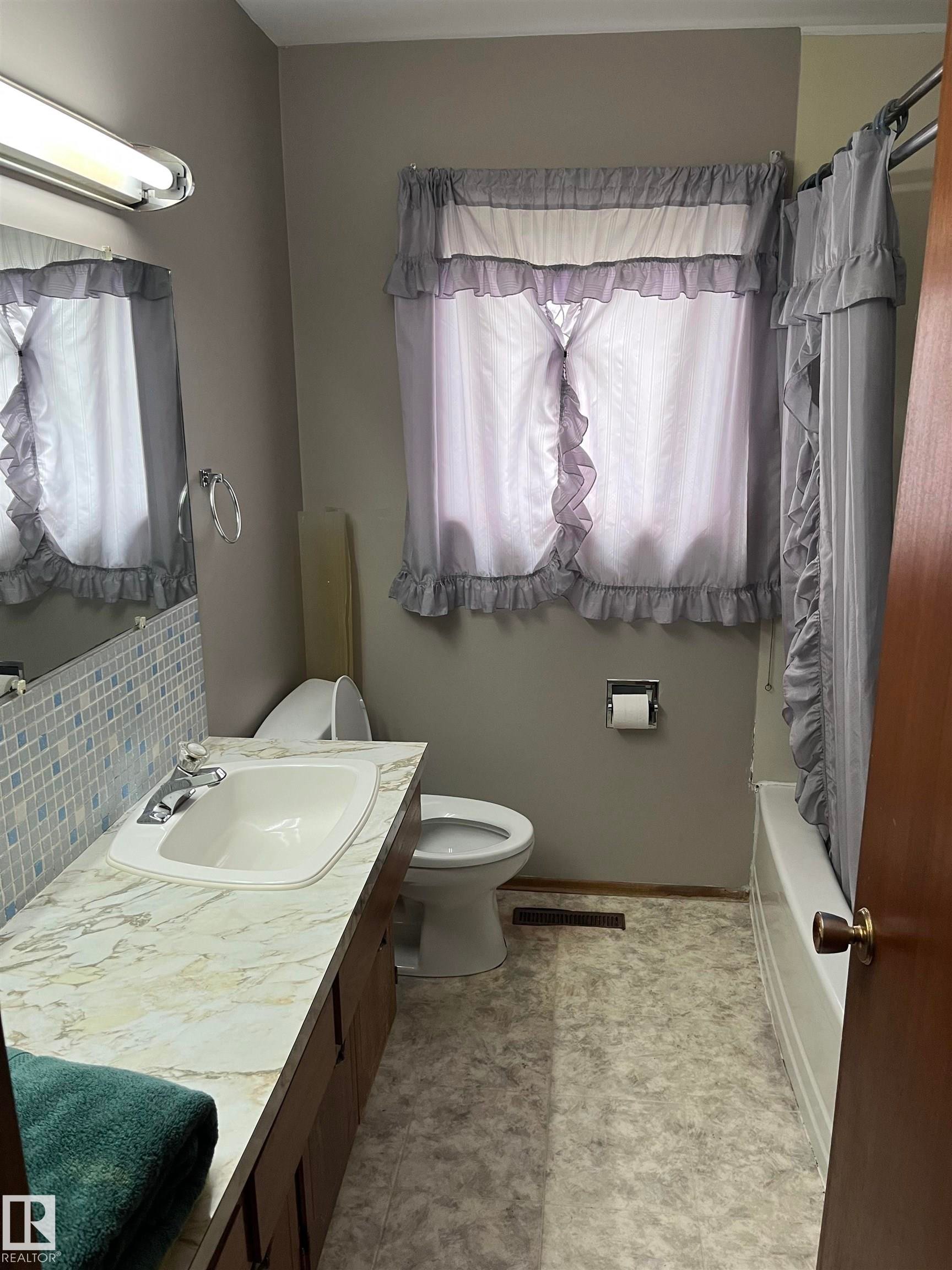Bathroom featuring a built-in vanity with a countertop and integrated sink, mosaic tile backsplash, and a window with privacy coverings - 5012 58 Avenue, Viking, AB - Indoor Photo Showing Bathroom
