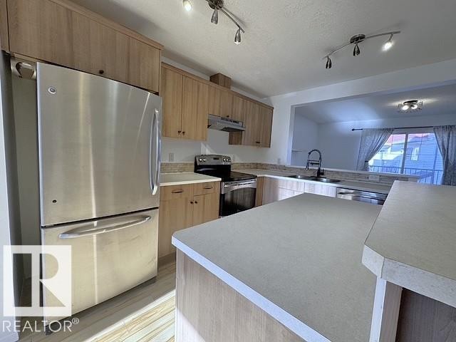 Modern kitchen featuring light wood-finish cabinetry, stainless steel appliances, light-toned countertops, and track lighting - #13 3751 12 Street, Edmonton, AB