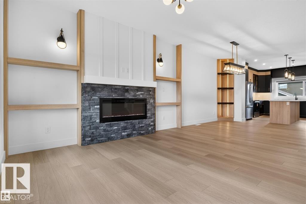 Living space featuring a linear electric fireplace with a stacked stone surround, wood-finish flooring, built-in shelving, sconce lighting, and an adjacent kitchen with an island and pendant lighting - 41 River Birch Way, St. Albert, AB