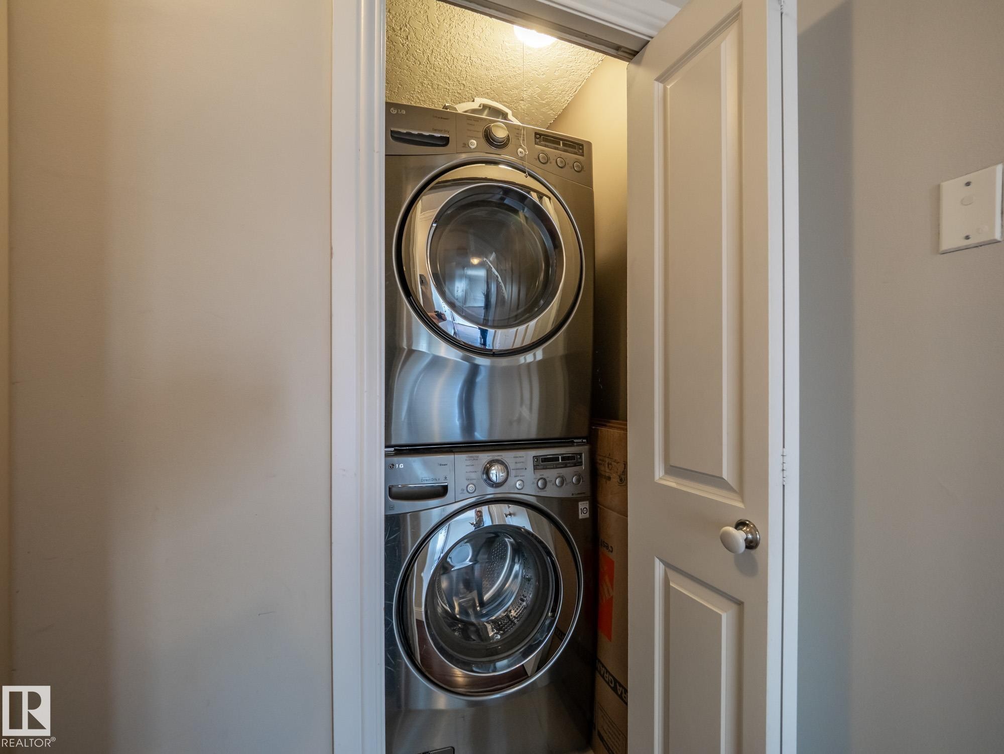 Stacked stainless steel washer and dryer units in a dedicated closet space, featuring a white paneled door and neutral wall tones - #Ph4 9725 106 St Nw, Edmonton, AB - Indoor Photo Showing Laundry Room