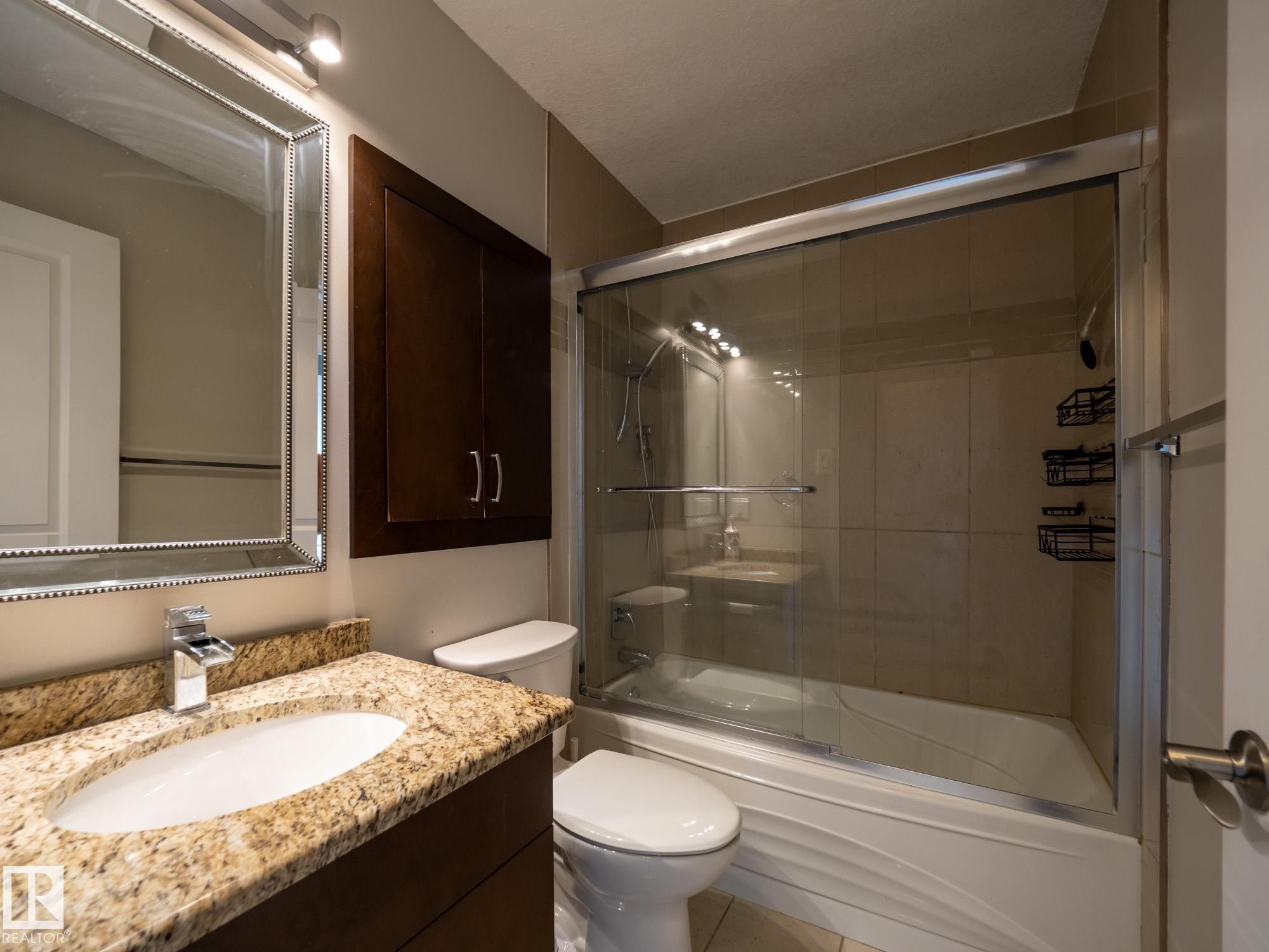 Bathroom featuring a granite-finish countertop with an undermount sink, a framed mirror with overhead lighting, a dark wood-finish wall cabinet, a white toilet, and a bathtub with a sliding glass enclosure - #Ph4 9725 106 St Nw, Edmonton, AB - Indoor Photo Showing Bathroom
