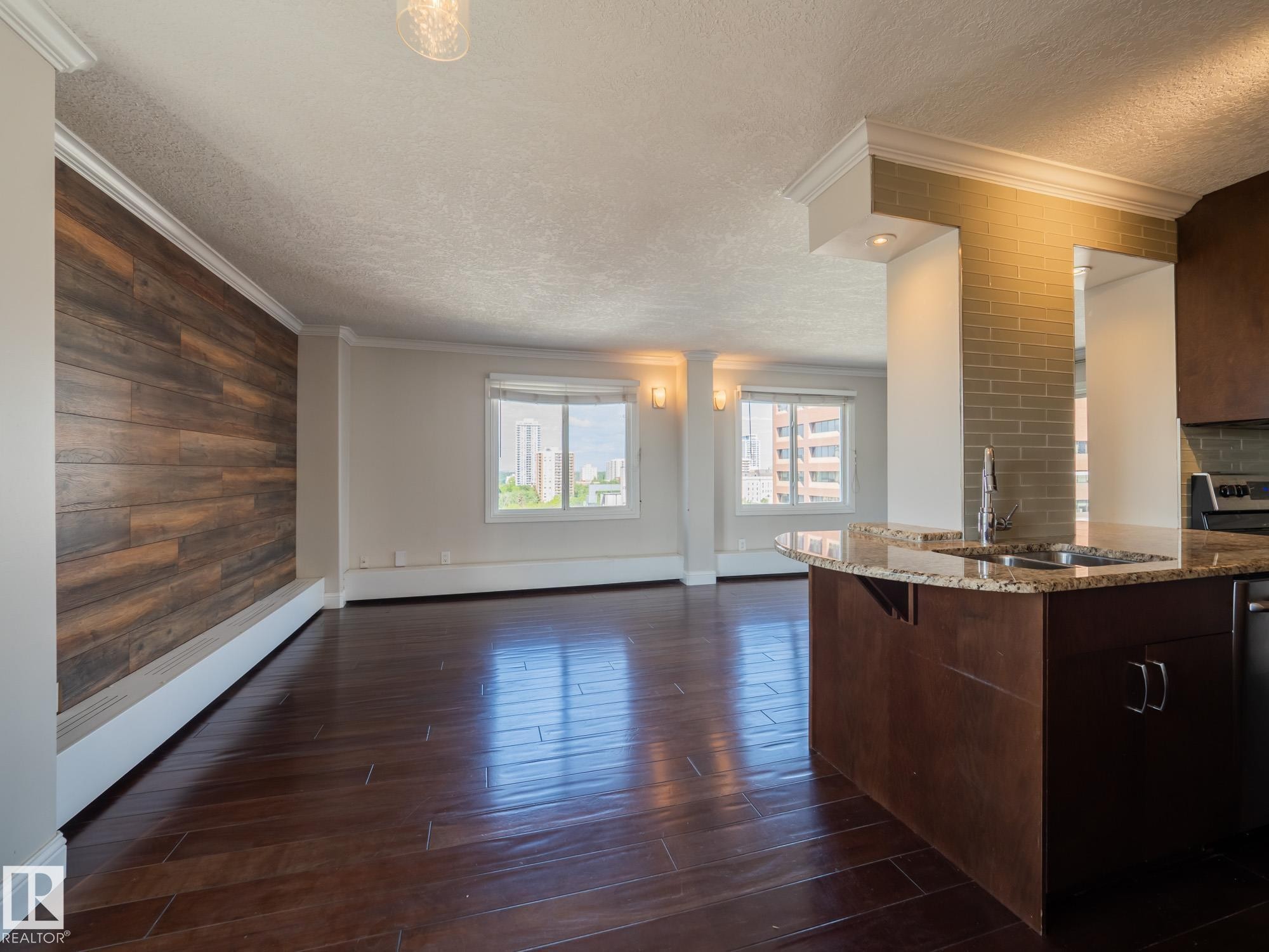 Open-concept living space featuring wood-finish flooring, a wood-paneled accent wall, and a kitchen island with a stone countertop and integrated sink - #Ph4 9725 106 St Nw, Edmonton, AB - Indoor