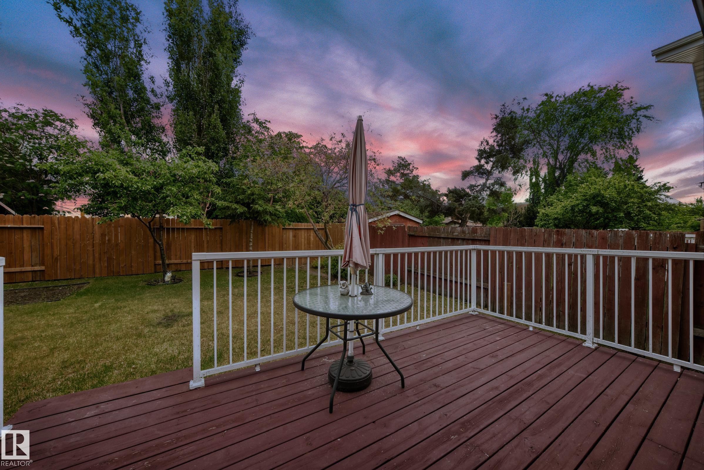 Expansive wood deck with white railings, offering direct access to a fenced backyard featuring a manicured lawn and mature trees - 3843 47 Street Nw, Edmonton, AB - Outdoor With Deck Patio Veranda