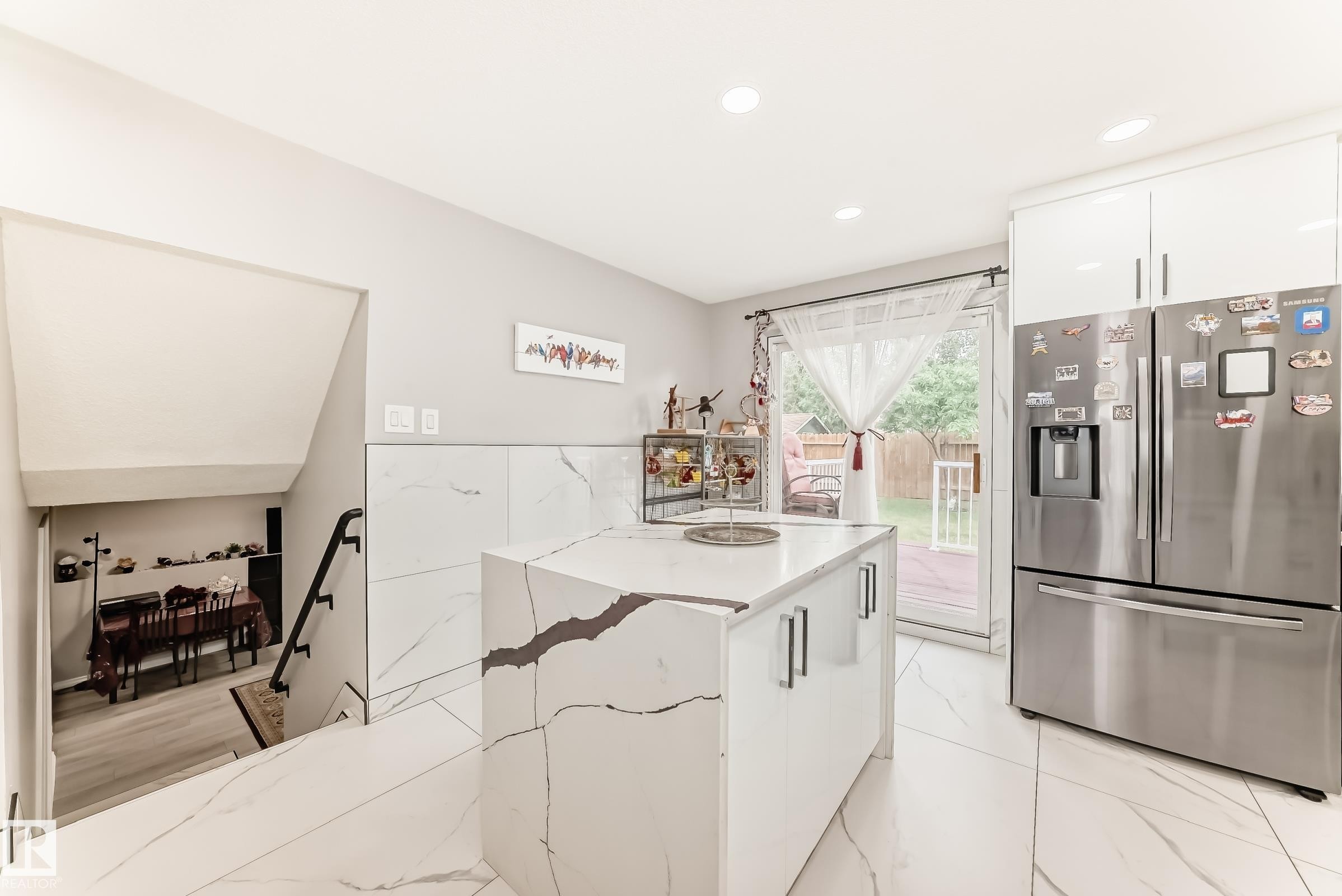 Contemporary kitchen featuring a central island with a waterfall countertop and integrated storage, stainless steel refrigerator, white gloss cabinetry, and large format porcelain tile flooring - 3843 47 Street Nw, Edmonton, AB - Indoor Photo Showing Kitchen