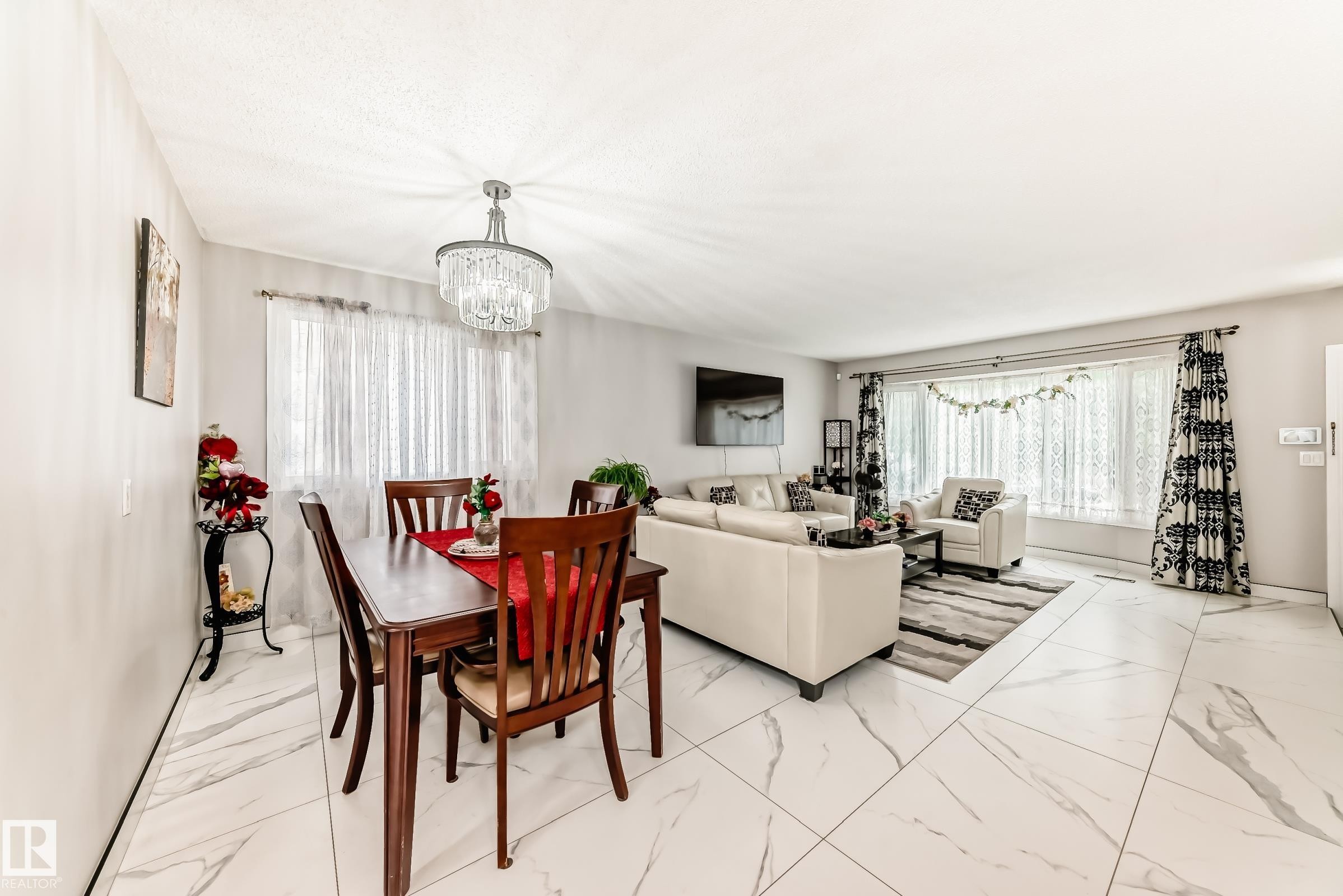Open-concept living and dining areas featuring large format white tile flooring, a crystal chandelier, and a wide window with sheer and patterned curtains - 3843 47 Street Nw, Edmonton, AB - Indoor
