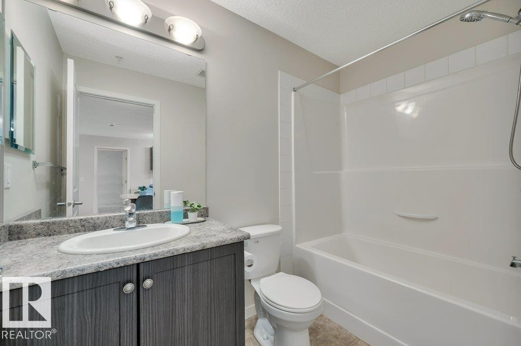 Full bathroom featuring a single vanity with wood-finish cabinetry, a white oval sink, and a speckled countertop - 219 1820 Rutherford Road, Edmonton, AB