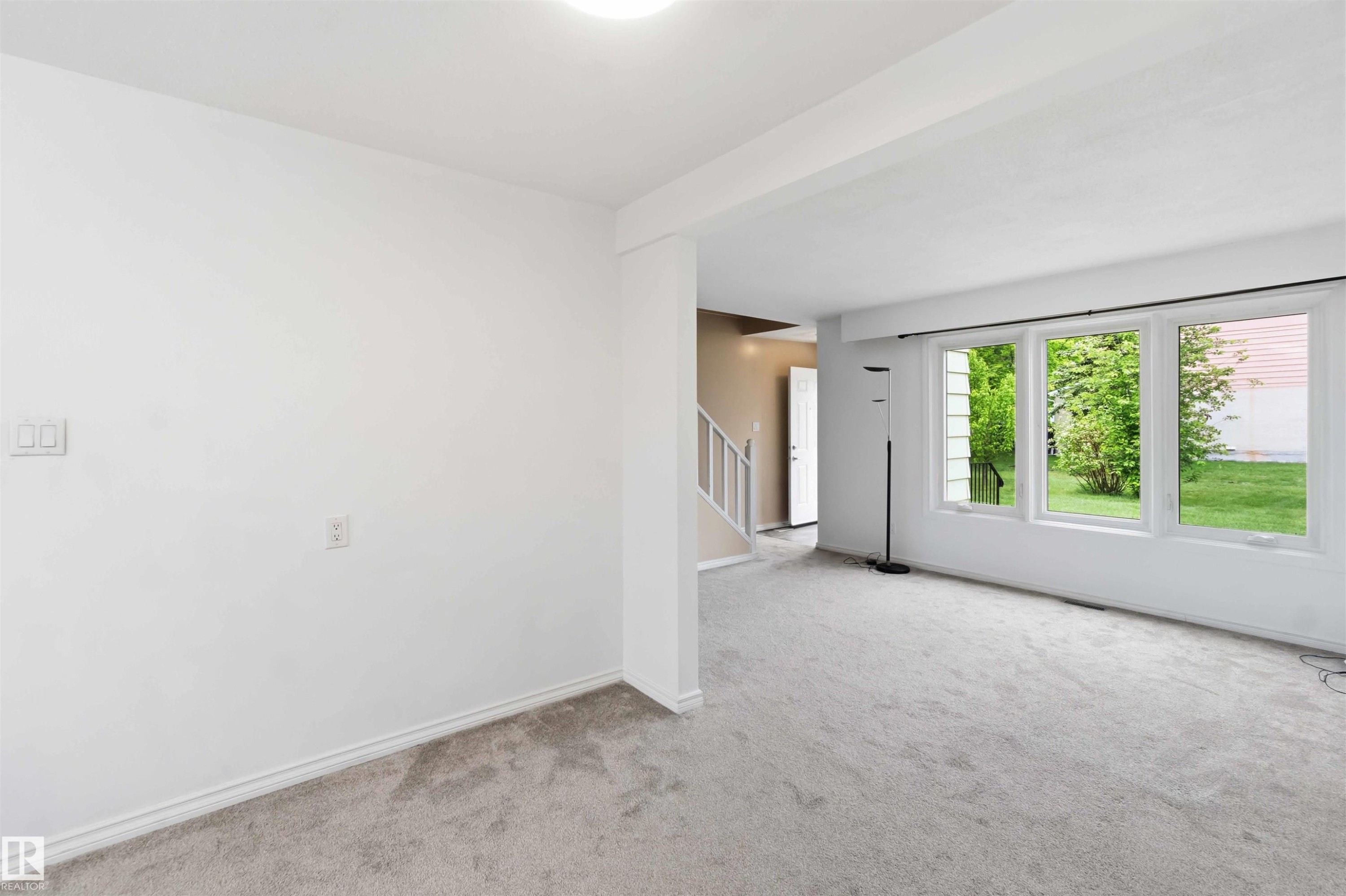 Spacious room featuring light gray carpeting, crisp white walls, multiple large windows, and a partially visible staircase with white railings - 17325 85 Avenue Nw, Edmonton, AB