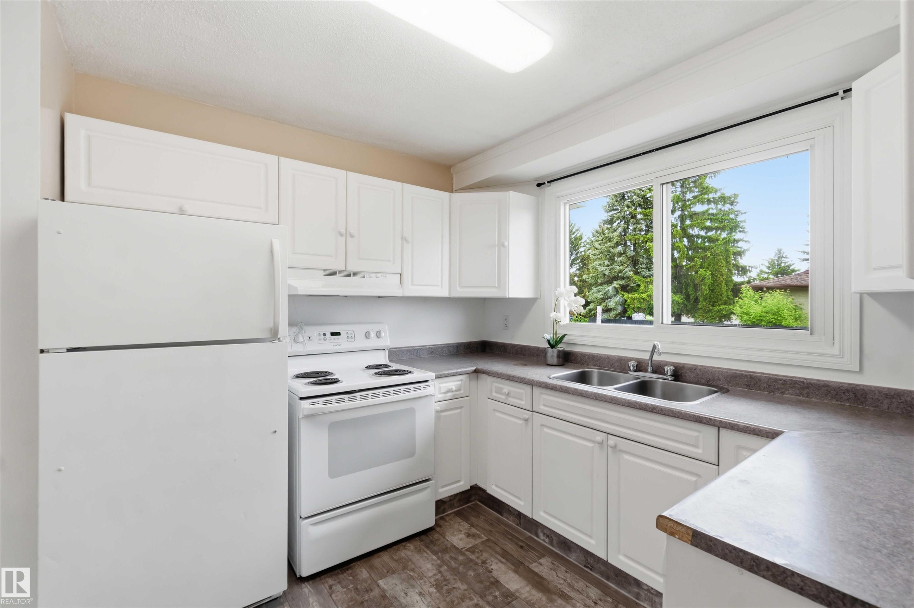 Bright kitchen featuring white cabinetry, a double basin stainless steel sink, wood-finish flooring, and a large window overlooking mature trees - 17325 85 Avenue Nw, Edmonton, AB